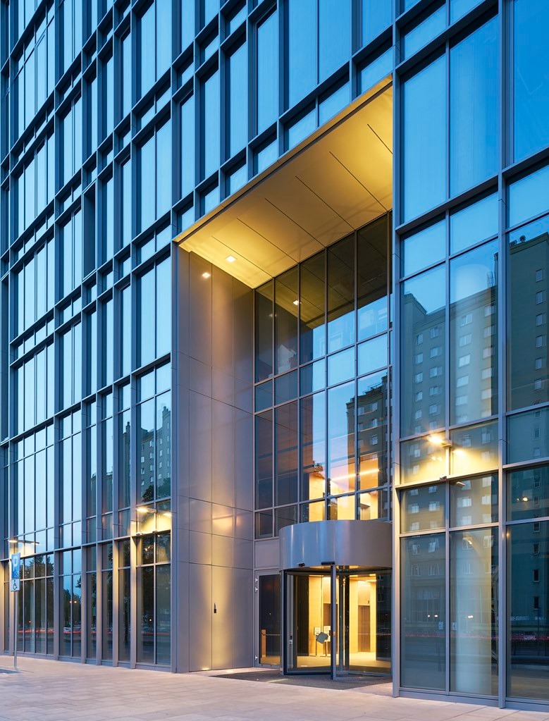 Fasada nowoczesnego biurowca z szklaną elewacją i obrotowymi drzwiami wejściowymi, oświetlona ciepłym światłem.