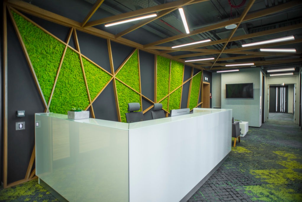 Nowoczesne biuro: biała lada recepcyjna na tle ściany z zielonego mchu, oświetlonej liniowymi lampami sufitowymi, z elementami drewnianej konstrukcji geometrycznej.