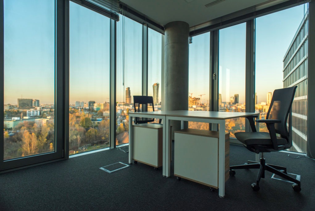 Nowoczesne biuro z widokiem na miasto o zachodzie słońca, minimalistyczne biurko z szafkami na kółkach i ergonomiczne krzesło biurowe, szara wykładzina dywanowa i betonowy filar.