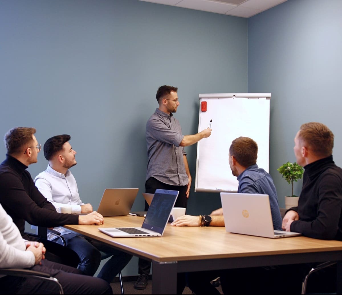 Mężczyzna w szarej koszuli prezentuje coś na białej tablicy flipchart grupie osób siedzących przy drewnianym stole z laptopami w biurze.