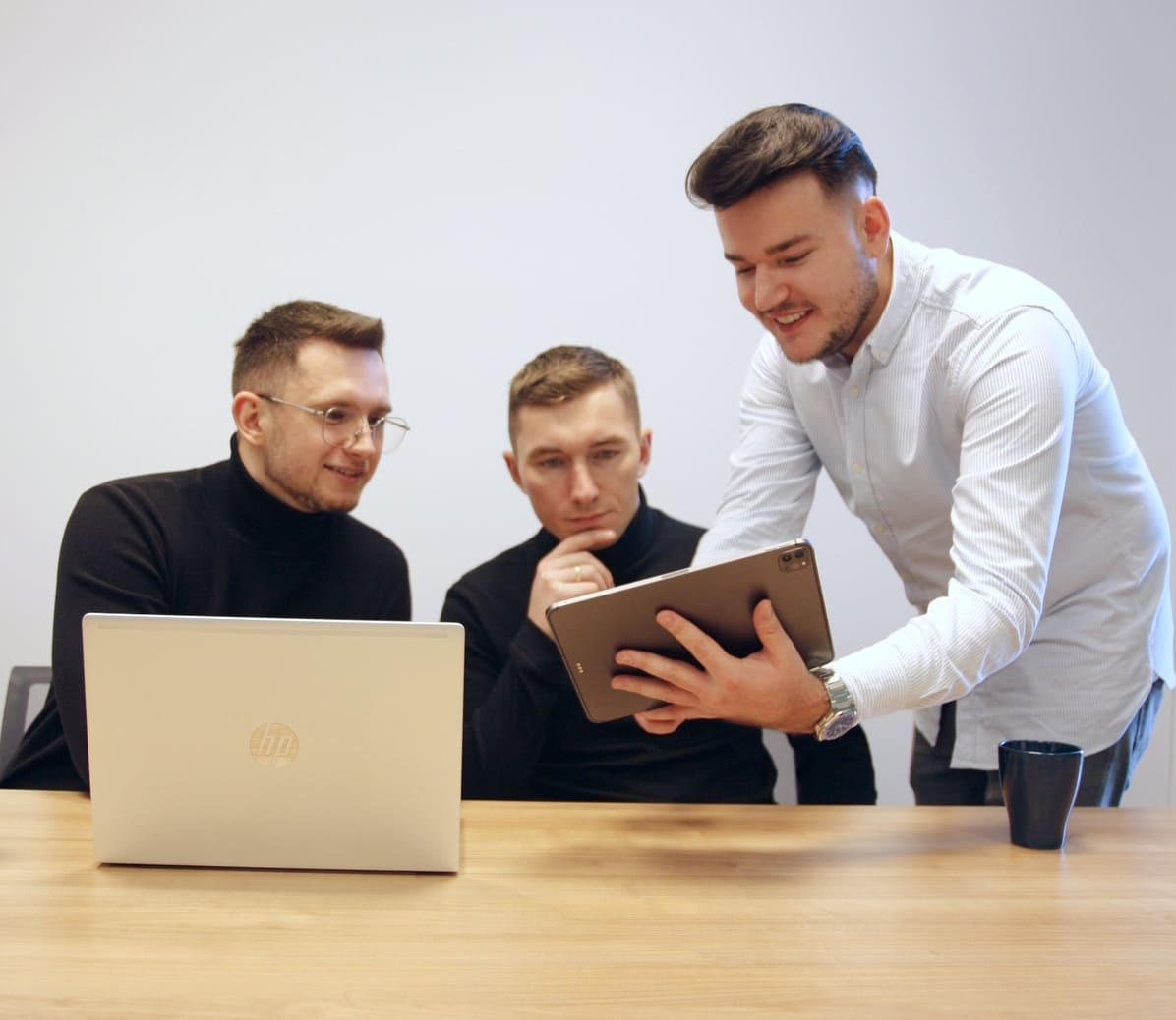 Trzech mężczyzn analizuje dane na laptopie HP i tablecie, siedząc przy jasnym drewnianym stole w minimalistycznym biurze.