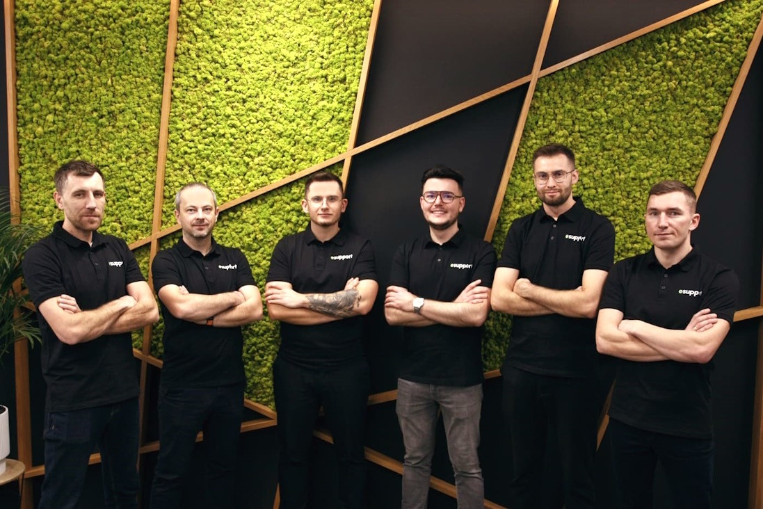 Zespół sześciu mężczyzn w czarnych koszulkach polo z logo firmy, stojących na tle ściany z mchu i geometrycznych drewnianych elementów dekoracyjnych.