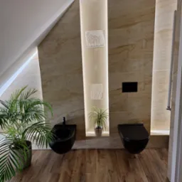 Nowoczesna łazienka na poddaszu z beżowymi płytkami imitującymi marmur, podświetlanymi niszami z dekoracjami i roślinami, oraz wiszącą, czarną armaturą.