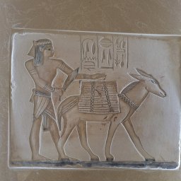 Batiment Greta Babinetz-Brzoza - Płaskorzeźba przedstawiająca Egipcjanina prowadzącego osła z towarem, z inskrypcjami hieroglificznymi powyżej, na beżowym tle.