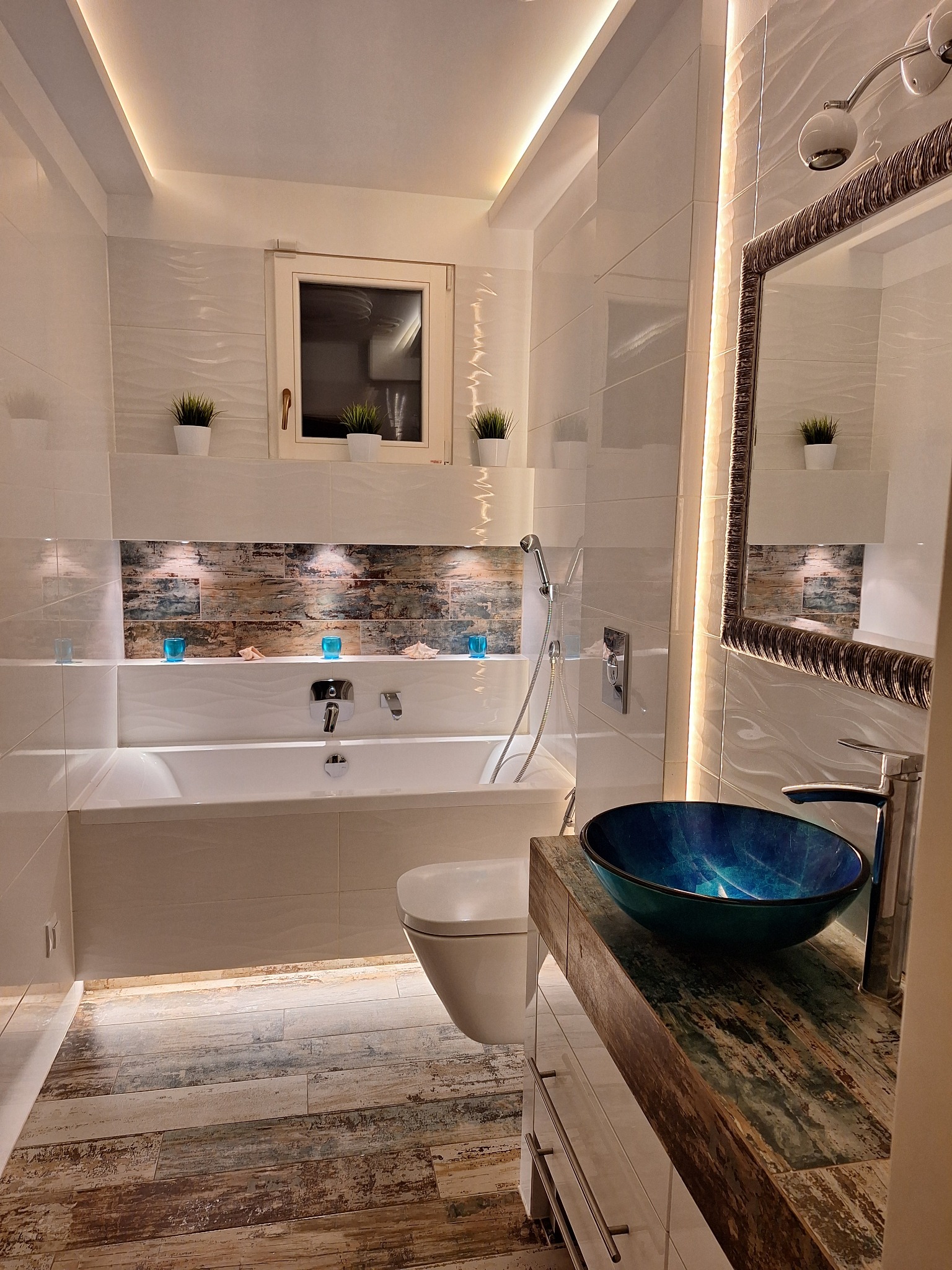 Nowoczesna łazienka z podświetlanymi półkami, wanną, toaletą i umywalką w kolorze morskim, drewnopodobną podłogą i lustrem w ozdobnej ramie.