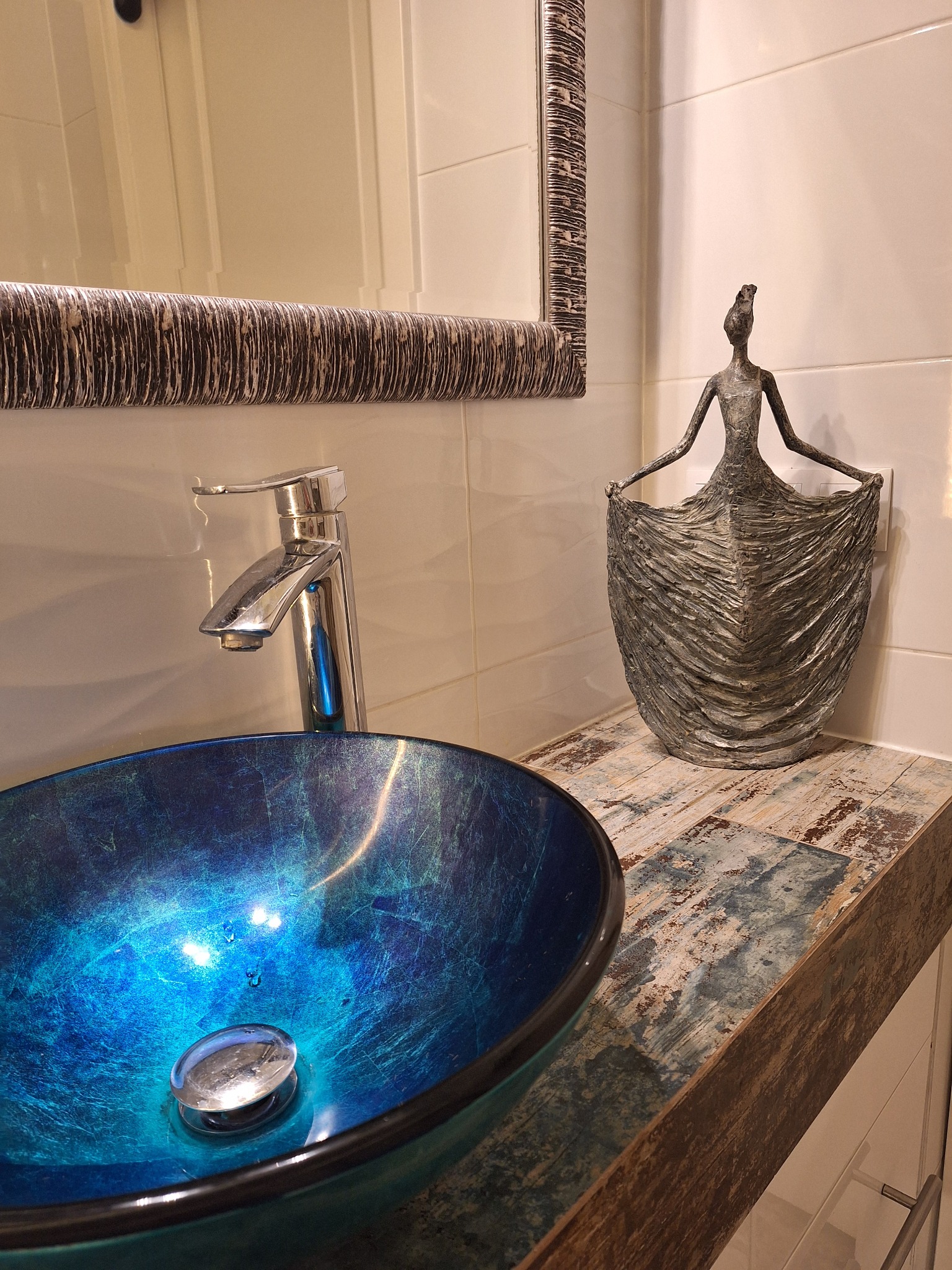 Błękitna, szklana umywalka na drewnianym blacie z metalową armaturą, obok dekoracyjna statuetka kobiety i lustro w ramie o fakturze kory drzewa na tle białych płytek.