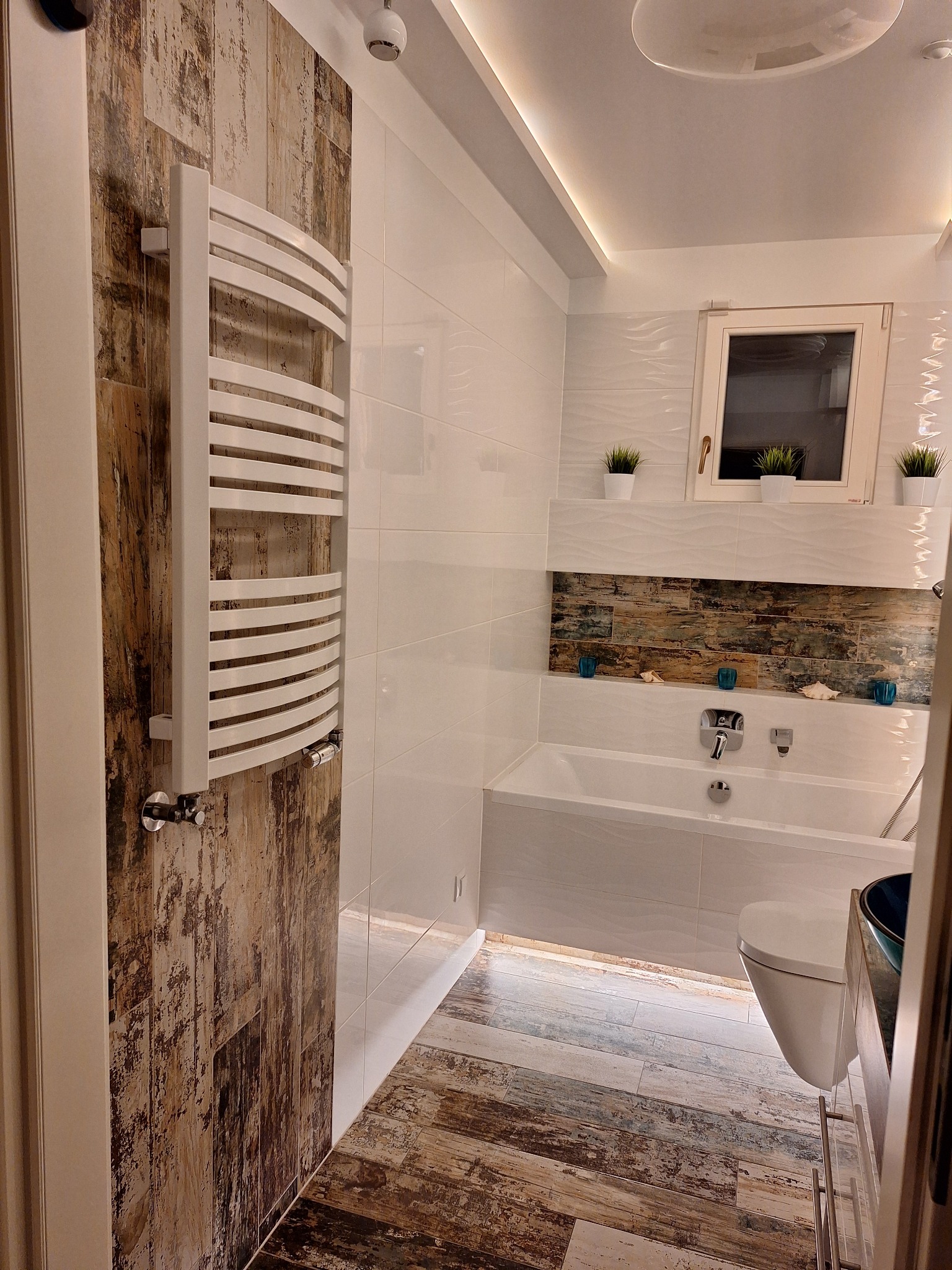 Nowoczesna łazienka z białymi płytkami, wanną, toaletą i grzejnikiem drabinkowym na ścianie z drewnopodobnych płytek, oświetlona listwami LED i lampą sufitową.