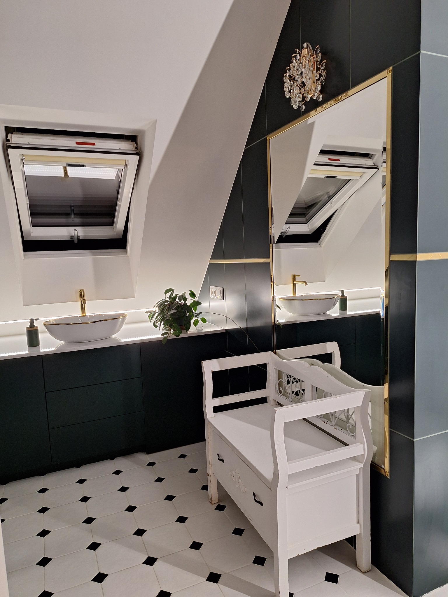 Nowoczesna łazienka na poddaszu z białymi umywalkami, złotą armaturą, czarno-białą podłogą w karo, lustrem w złotej ramie i białą ławeczką z ornamentami.