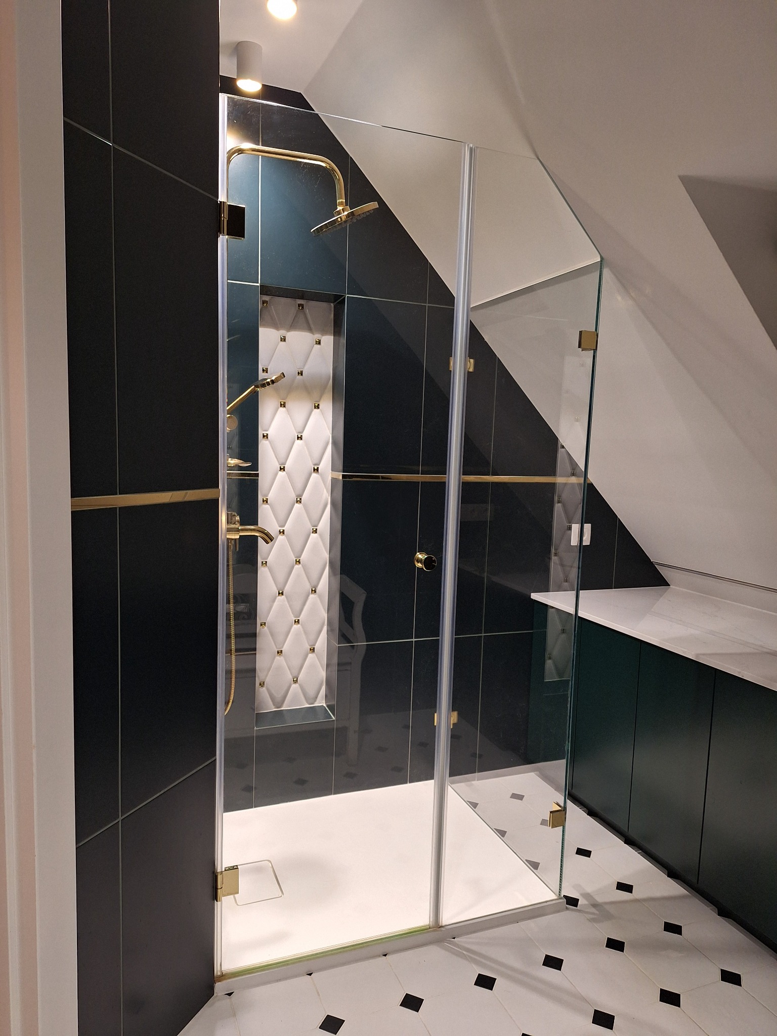 Nowoczesna kabina prysznicowa z czarnymi płytkami, złotymi elementami i dekoracyjną ścianą w romby w łazience na poddaszu.