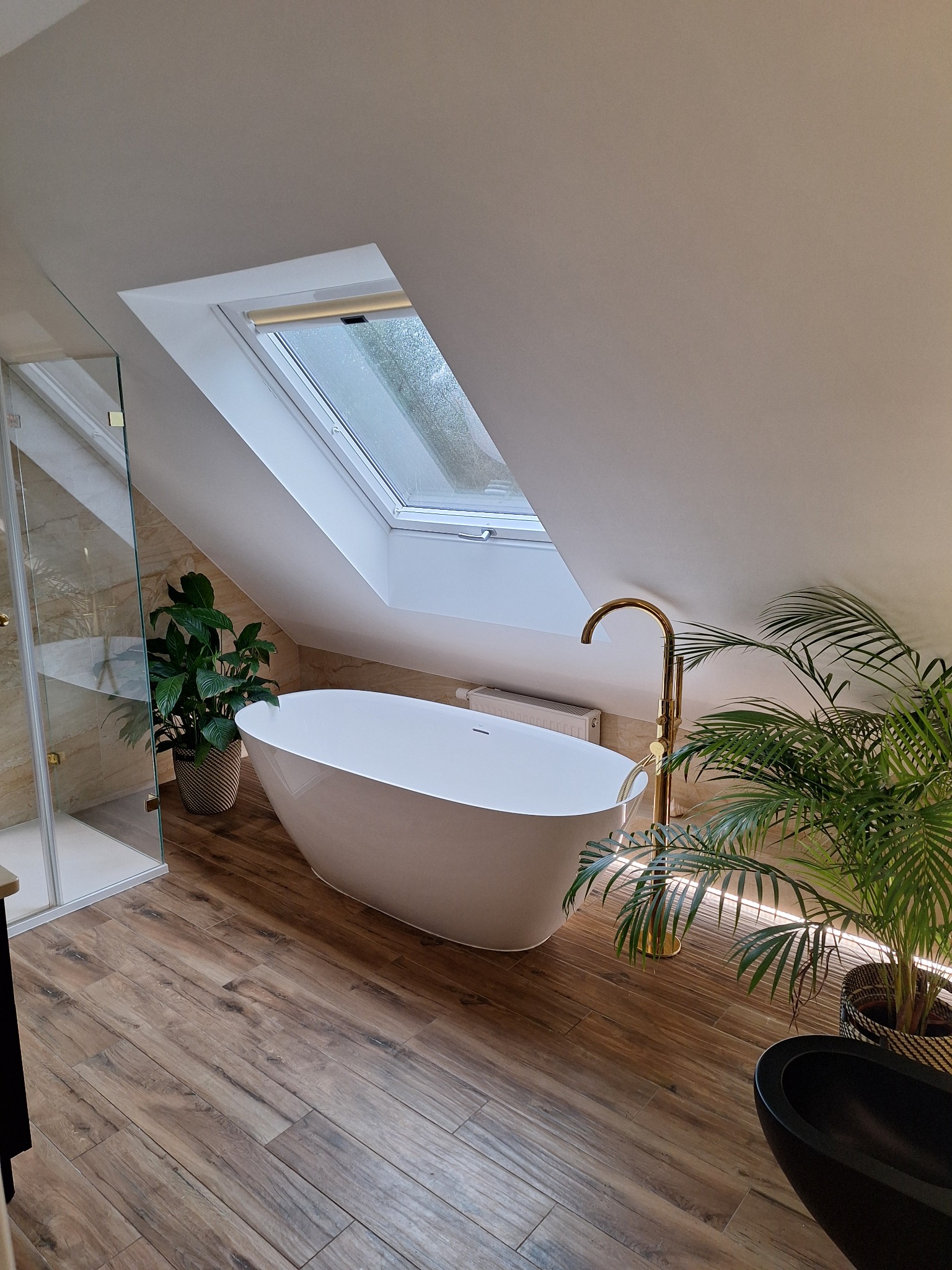 Nowoczesna łazienka na poddaszu z wolnostojącą białą wanną, złotą armaturą, roślinami doniczkowymi i kabiną prysznicową ze złotymi detalami.