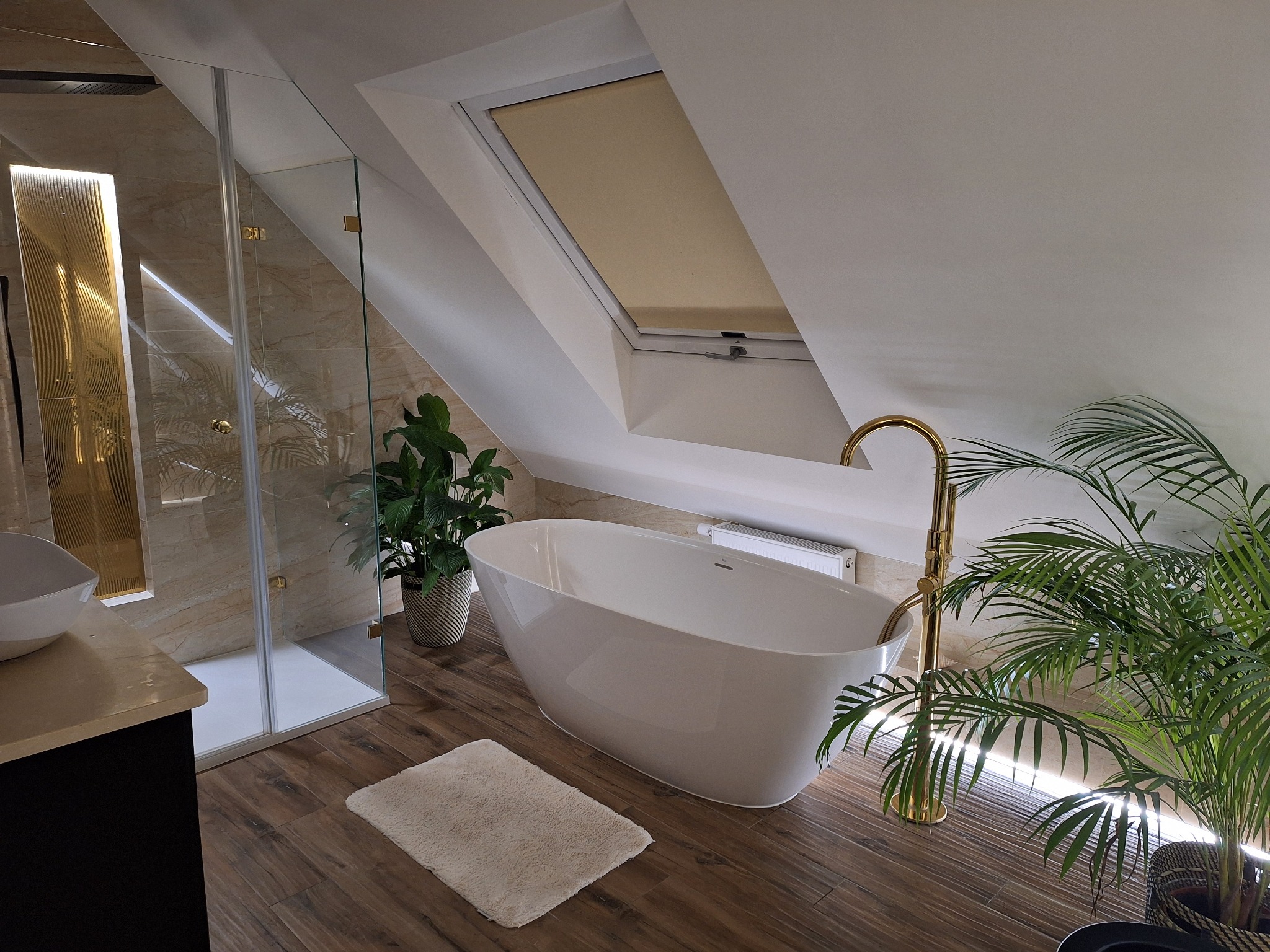 Nowoczesna łazienka na poddaszu z wolnostojącą wanną, złotą armaturą, kabiną prysznicową typu walk-in i roślinami doniczkowymi.