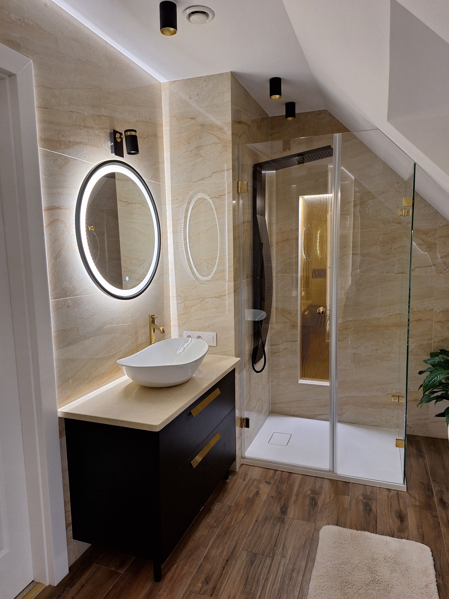 Nowoczesna łazienka na poddaszu z beżowymi płytkami, okrągłym lustrem z podświetleniem LED, białą umywalką nablatową, czarną szafką z złotymi uchwytami i kabiną prysznicową ze złotymi akcentami.
