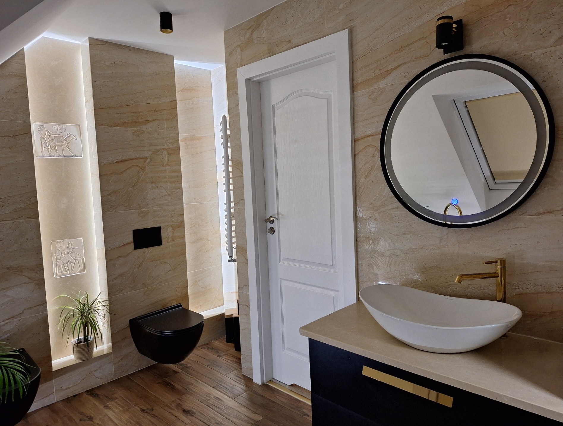 Nowoczesna łazienka w beżowych odcieniach z wiszącą czarną toaletą, okrągłym lustrem podświetlanym LED i umywalką nablatową z złotą armaturą. Na ścianie dekoracyjny relief z motywem egipskim.