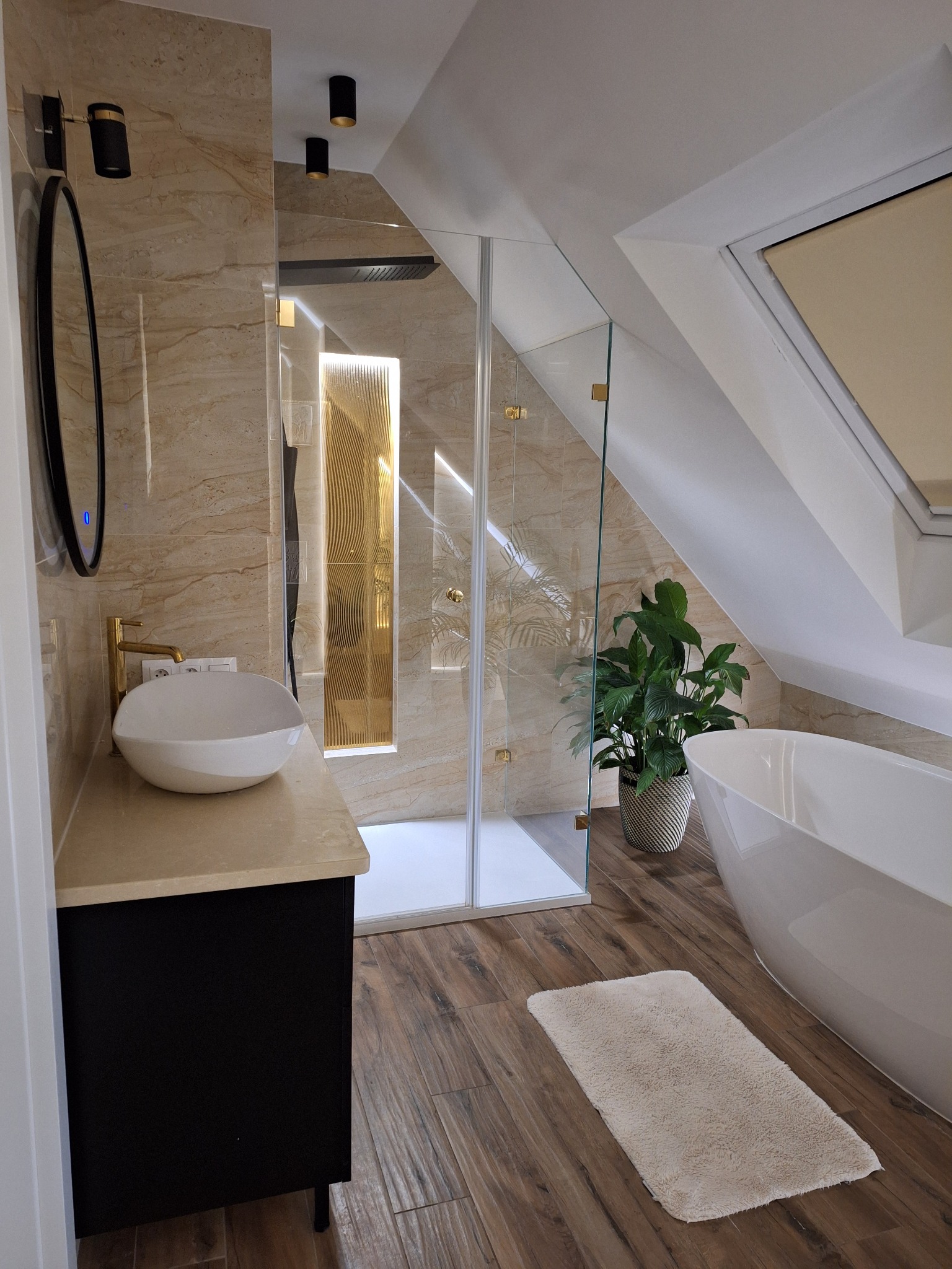 Jasna łazienka na poddaszu z kabiną prysznicową ze złotymi zdobieniami, wolnostojącą wanną i umywalką nablatową na czarnej szafce.