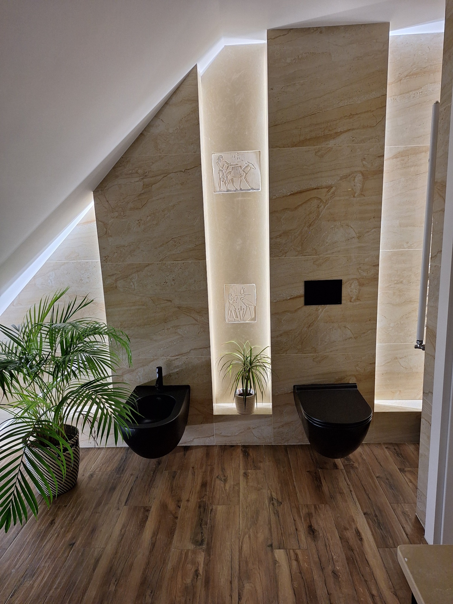 Nowoczesna łazienka na poddaszu z beżowymi płytkami imitującymi marmur, podświetlanymi niszami z dekoracjami i roślinami, oraz wiszącą, czarną armaturą.