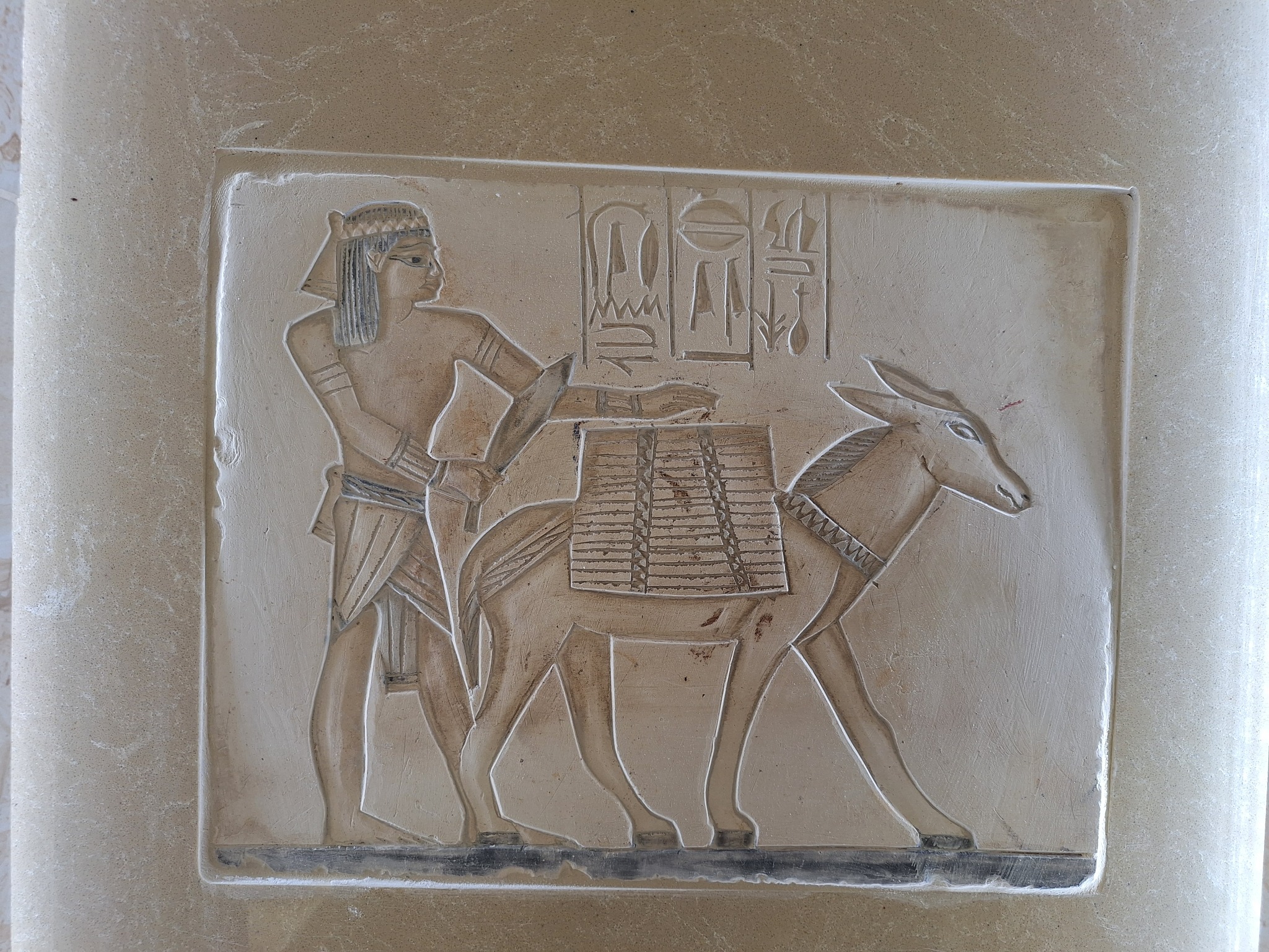 Płaskorzeźba przedstawiająca Egipcjanina prowadzącego osła z towarem, z inskrypcjami hieroglificznymi powyżej, na beżowym tle.