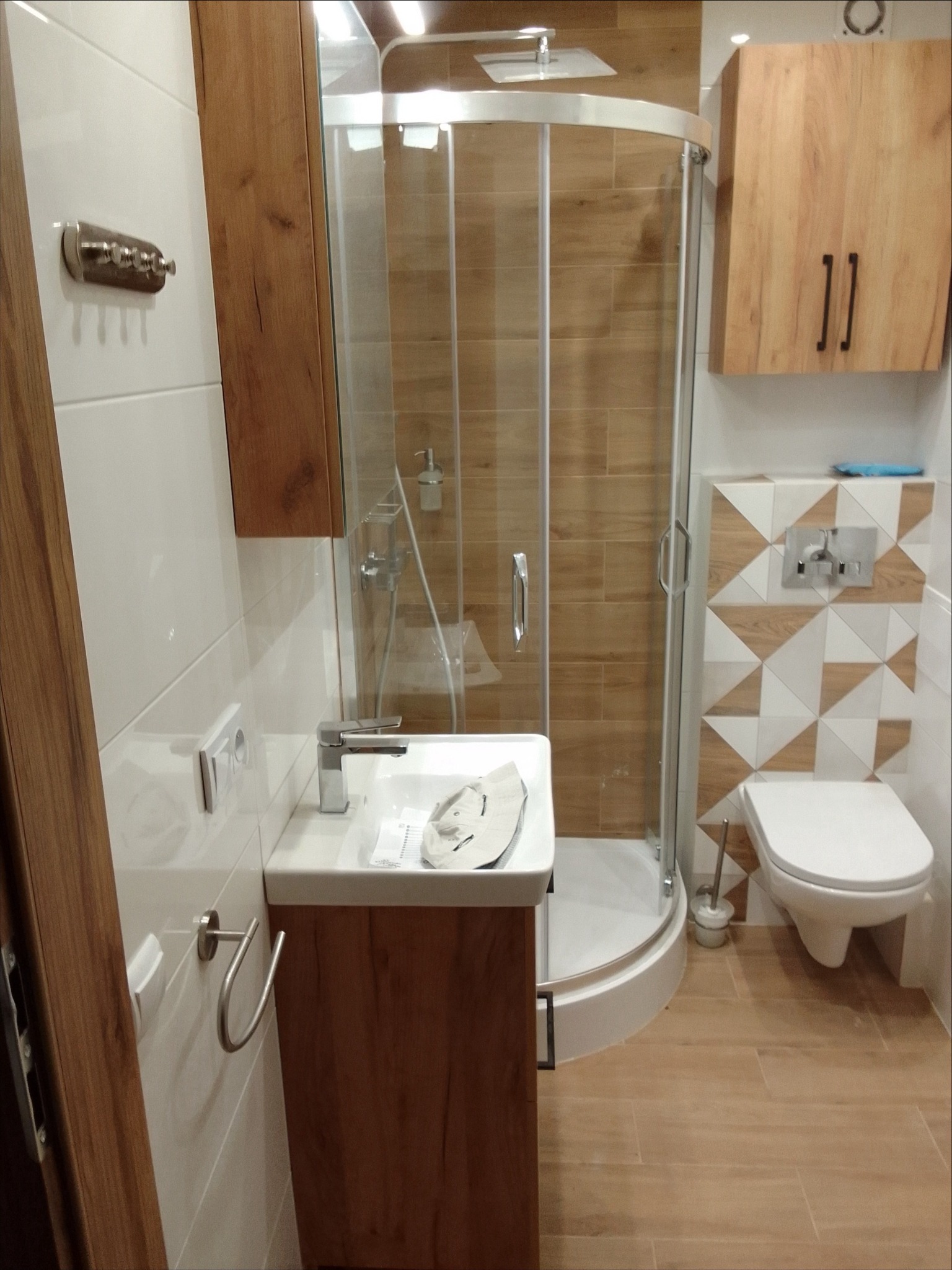 Nowoczesna łazienka z kabiną prysznicową, umywalką z szafką w kolorze drewna, toaletą i geometrycznymi płytkami na ścianie, całość utrzymana w ciepłej kolorystyce.