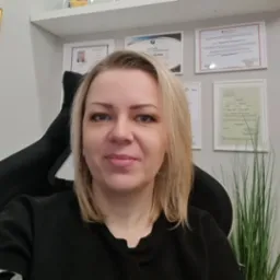 Portret kobiety z blond włosami, na ścianie za nią wiszą certyfikaty i dyplomy w ramkach, obok doniczka z zieloną rośliną.