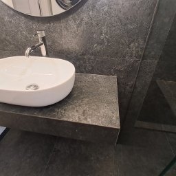 Robert Wawerzeniecki - Nowoczesna łazienka: biała umywalka nablatowa na szarej, kamiennej płycie, ściana i podłoga wyłożone ciemnymi płytkami imitującymi kamień, widoczny fragment kabiny prysznicowej.