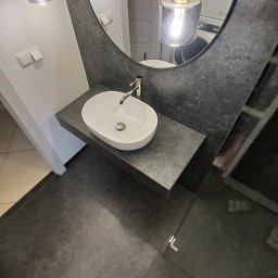 Robert Wawerzeniecki - Nowoczesna łazienka z owalną umywalką nablatową, szarą płytką na ścianie i podłodze oraz okrągłym lustrem z wiszącymi lampami. W tle widoczna pralka.