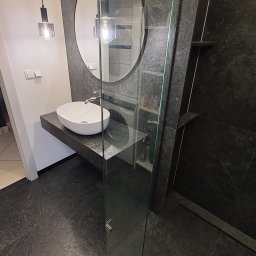 Robert Wawerzeniecki - Nowoczesna łazienka z owalną umywalką na blacie z ciemnego kamienia, okrągłe lustro i szklana kabina prysznicowa. Minimalistyczny design, ciemna kolorystyka.