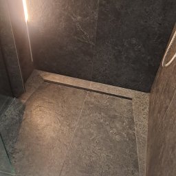 Robert Wawerzeniecki - Nowoczesna łazienka z szarymi płytkami imitującymi kamień, czarnym odpływem liniowym i podświetlaną półką. Minimalistyczny design, eleganckie wykończenie.