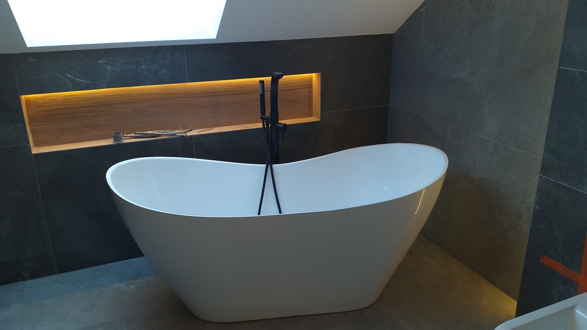 Wolnostojąca wanna w nowoczesnej łazience z szarymi płytkami imitującymi kamień, podświetlana wnęka w ścianie i czarna armatura.