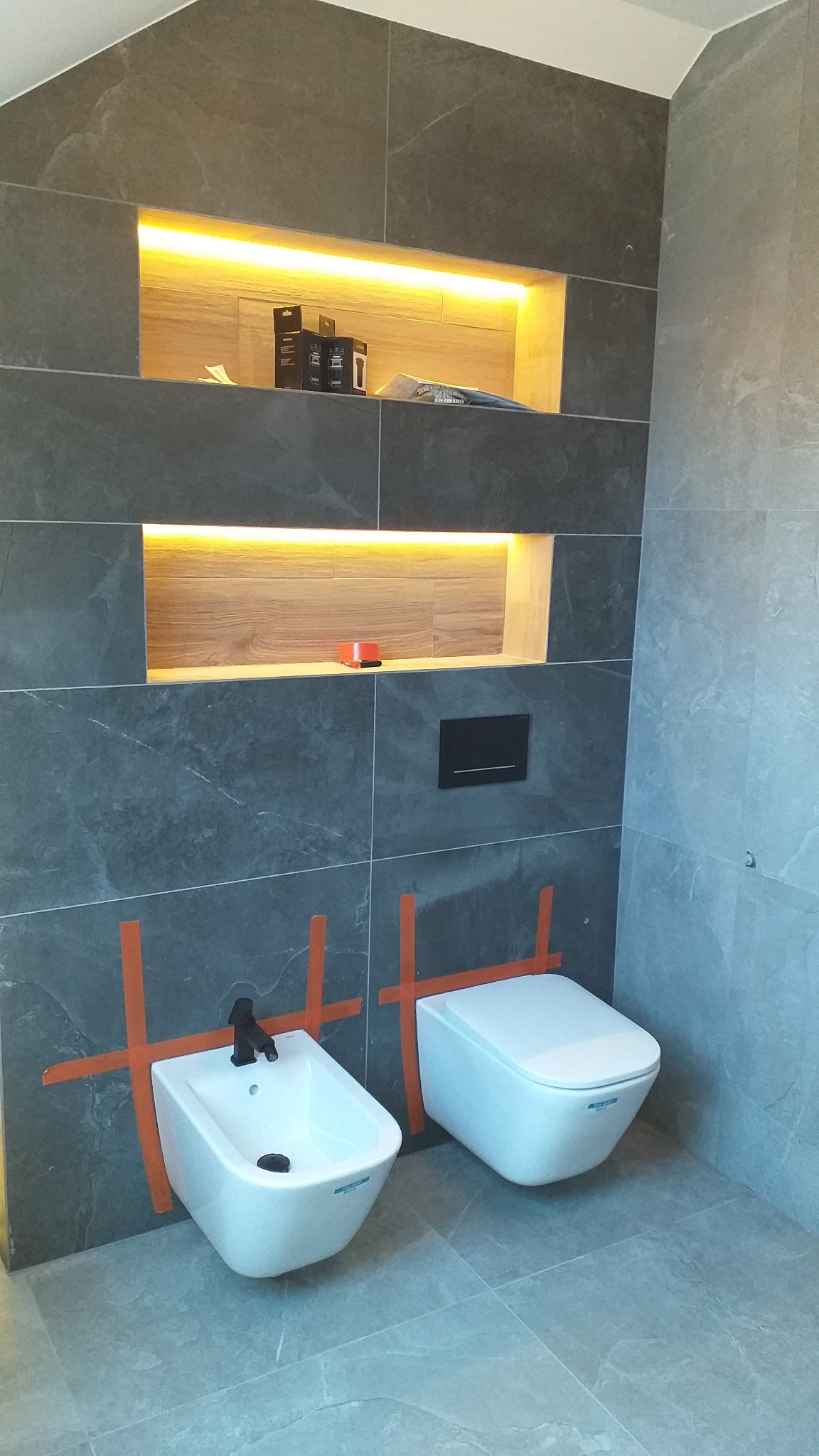 Nowoczesna łazienka z szarymi płytkami imitującymi kamień, podwieszany bidet z czarną armaturą i toaleta, obrysowane pomarańczową taśmą malarską. W ścianie wnęki z podświetleniem LED, na półce...