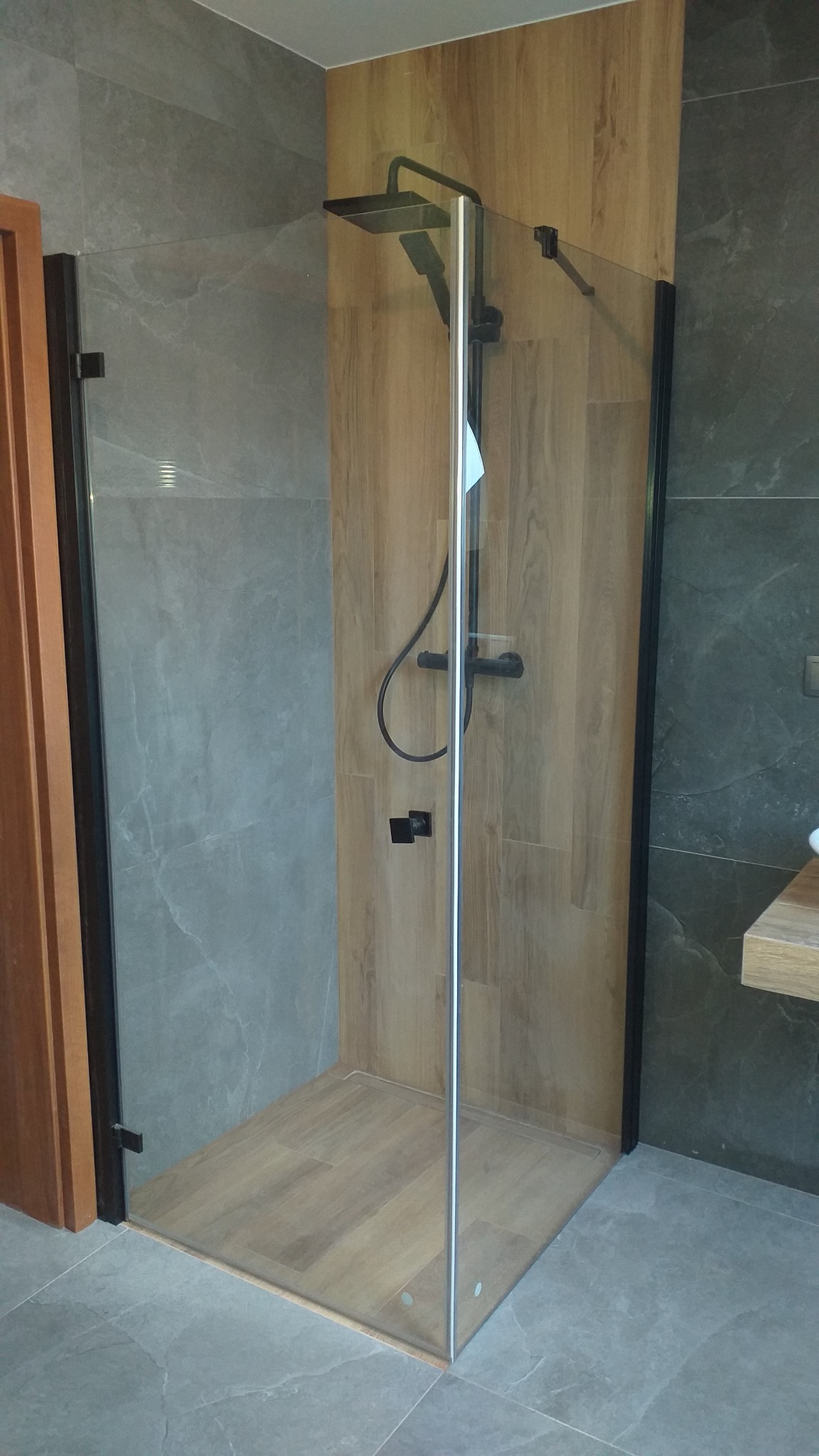 Nowoczesna kabina prysznicowa walk-in z czarną armaturą i drewnopodobnymi płytkami na ścianach i podłodze, widok z boku na szarą, marmurkową płytkę podłogową i częściowo widoczne drzwi wejściowe.