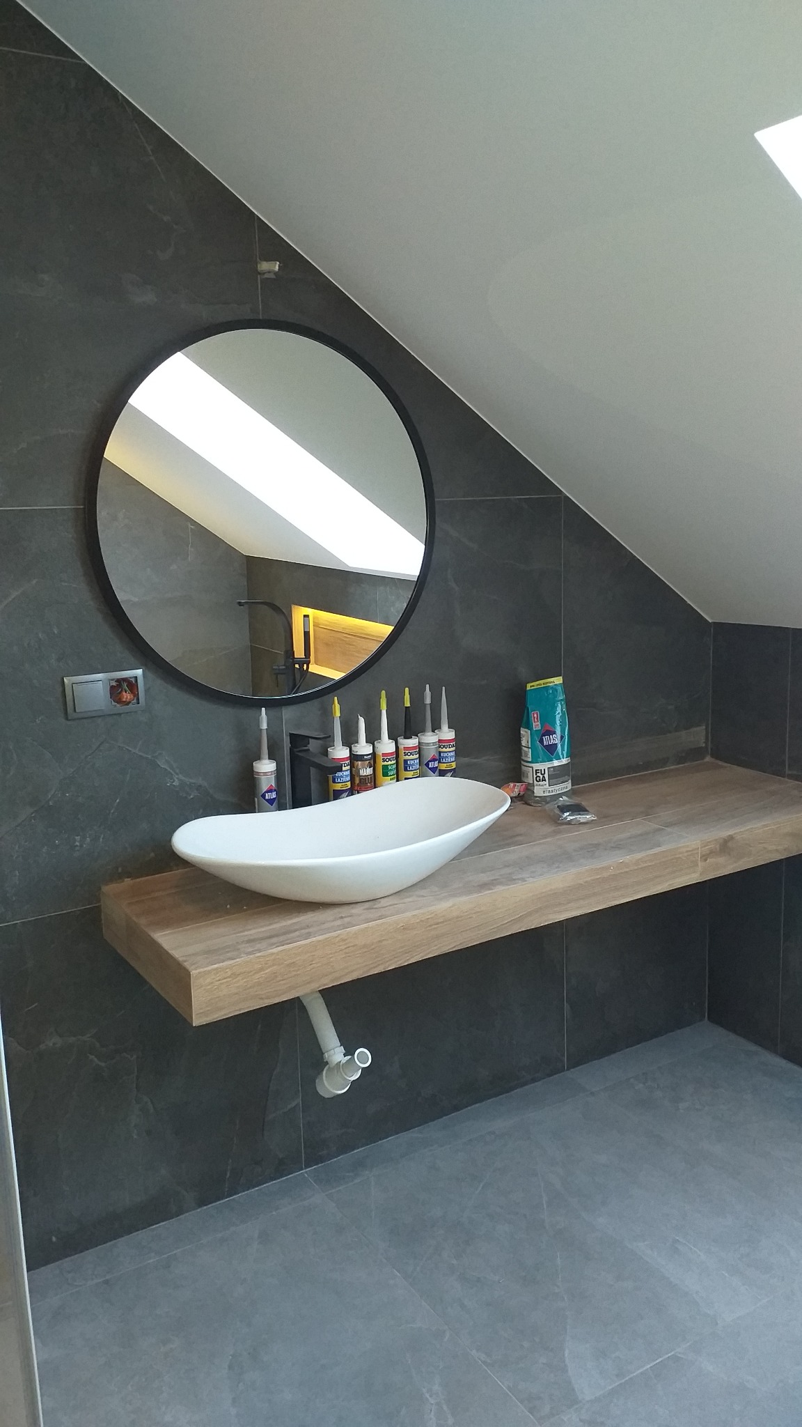 Nowoczesna łazienka na poddaszu z owalną umywalką nablatową, drewnianym blatem i okrągłym lustrem, widoczne tuby silikonu i opakowanie fugi.