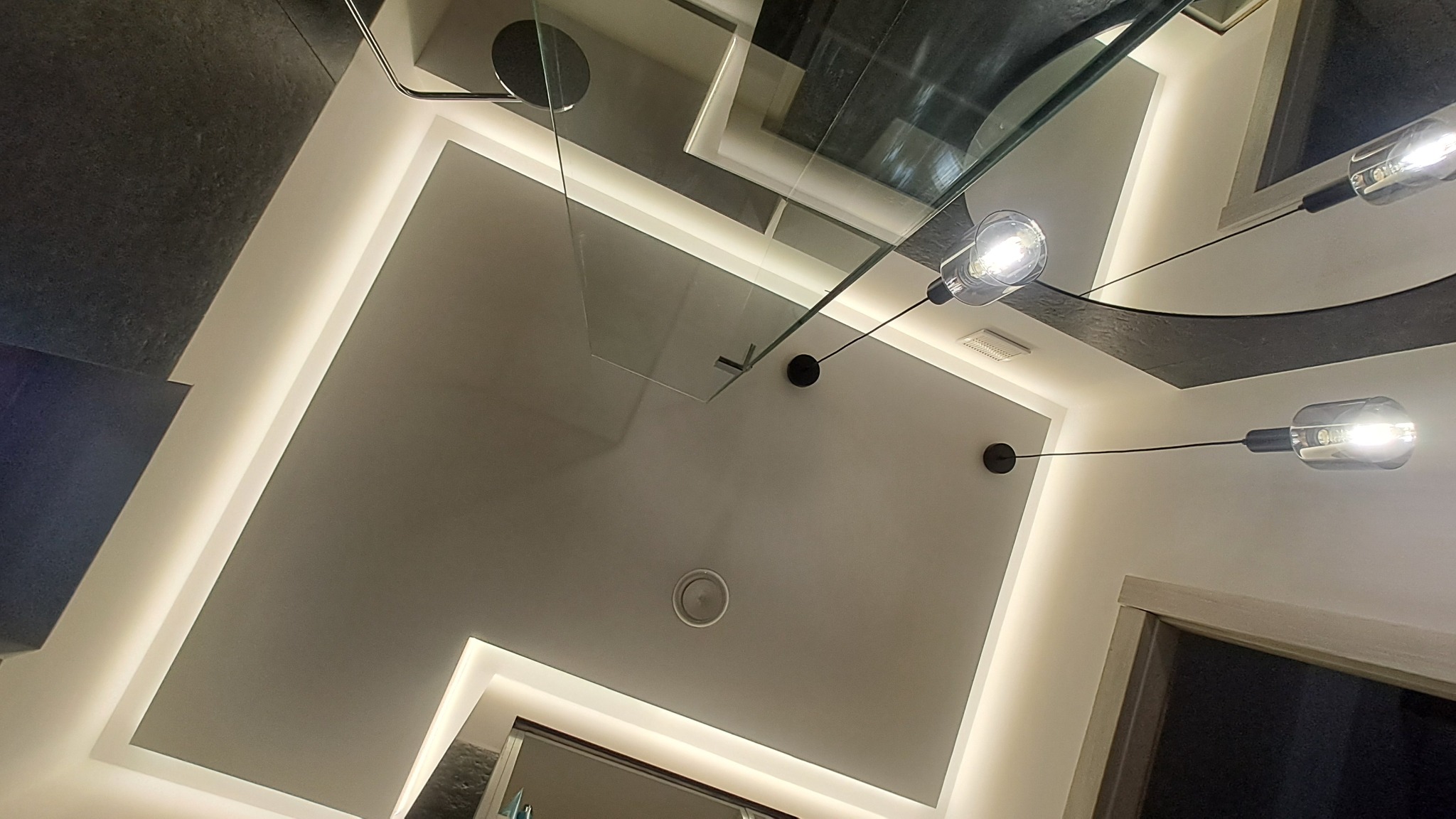 Nowoczesna łazienka z podświetlanym sufitem, szklanym panelem prysznicowym i designerskimi lampami wiszącymi. Minimalistyczny design z akcentami światła.