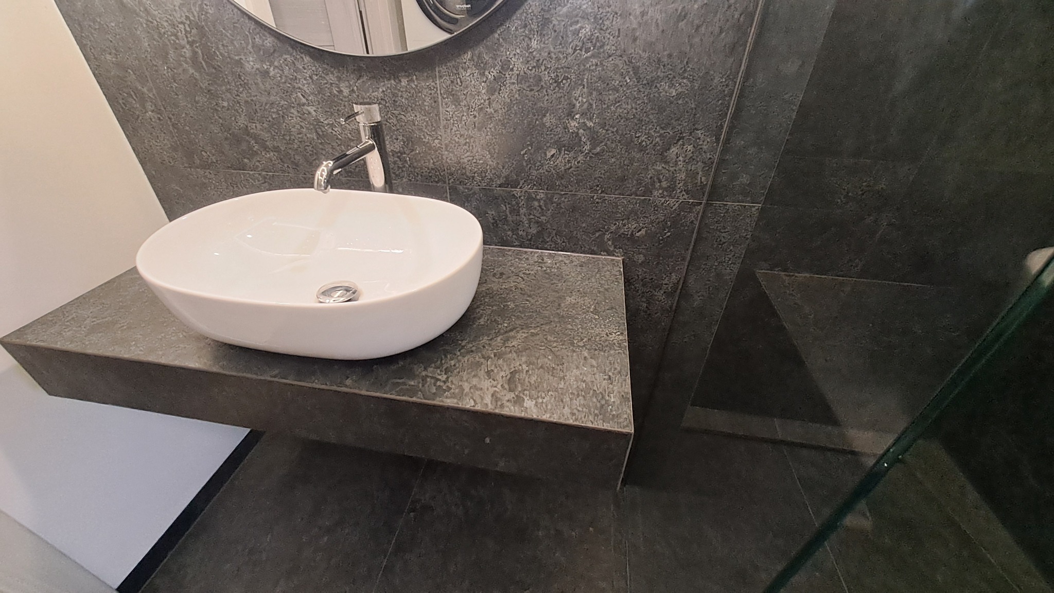 Nowoczesna łazienka: biała umywalka nablatowa na szarej, kamiennej płycie, ściana i podłoga wyłożone ciemnymi płytkami imitującymi kamień, widoczny fragment kabiny prysznicowej.