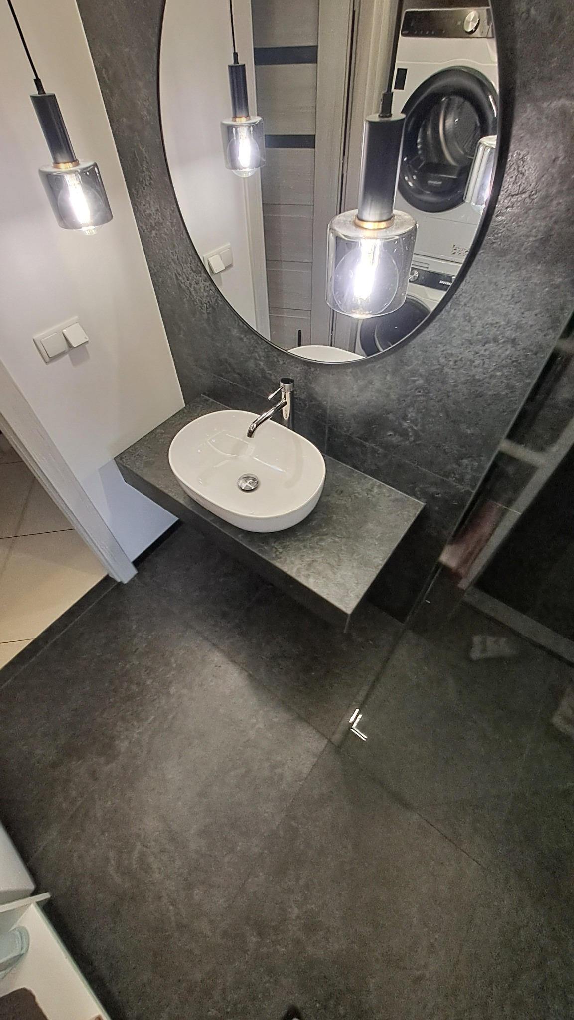 Nowoczesna łazienka z owalną umywalką nablatową, szarą płytką na ścianie i podłodze oraz okrągłym lustrem z wiszącymi lampami. W tle widoczna pralka.