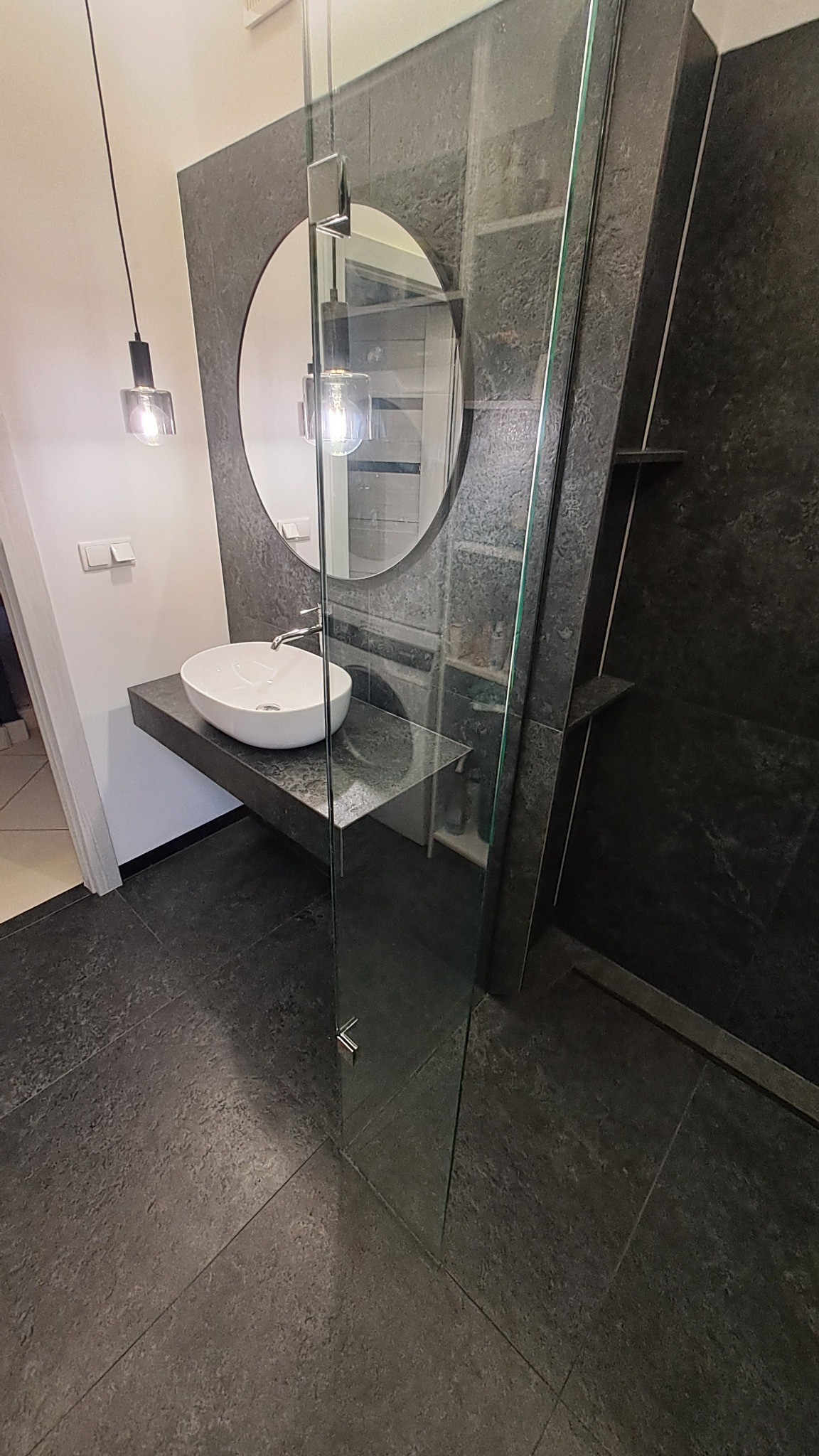 Nowoczesna łazienka z owalną umywalką na blacie z ciemnego kamienia, okrągłe lustro i szklana kabina prysznicowa. Minimalistyczny design, ciemna kolorystyka.