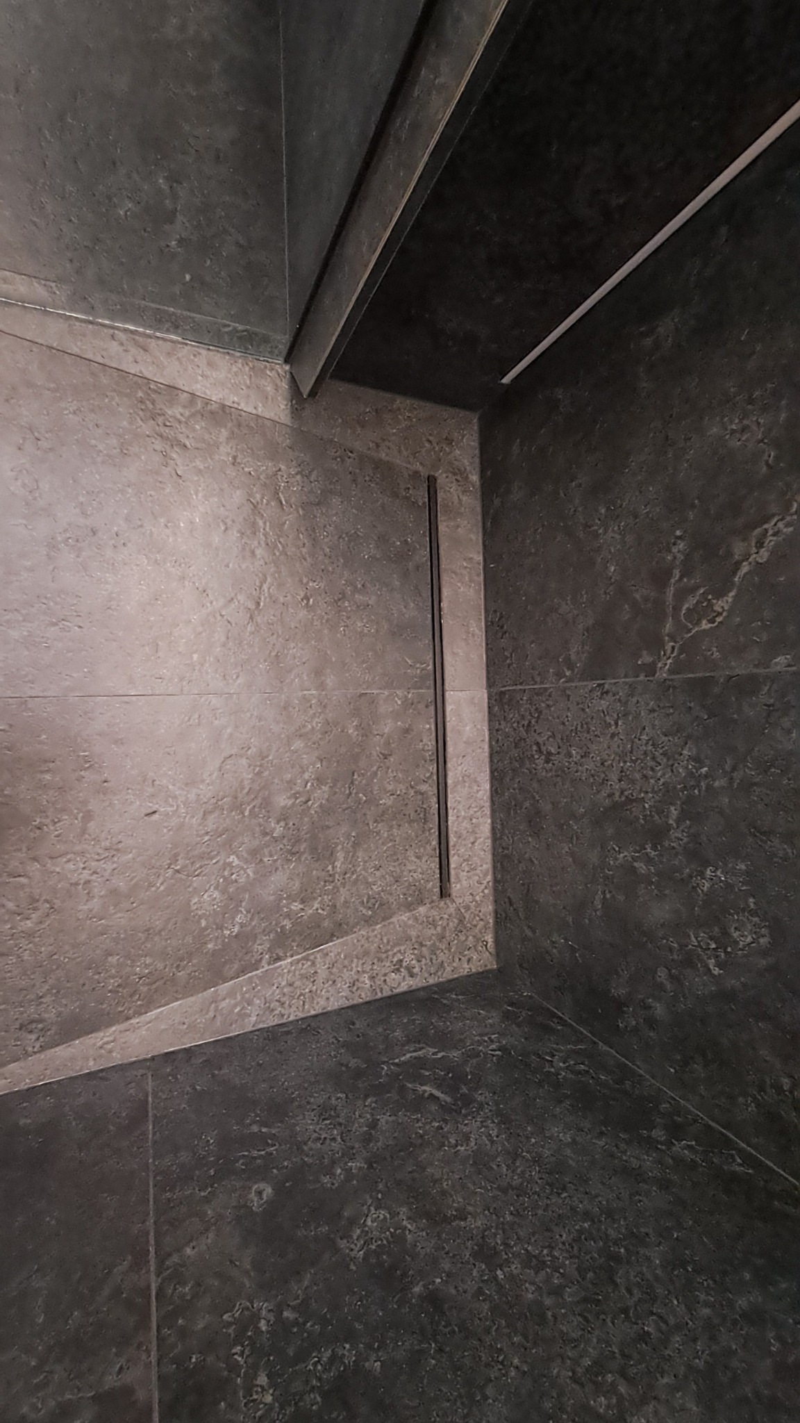 Nowoczesna kabina prysznicowa z odpływem liniowym i ciemnymi płytkami imitującymi kamień. Minimalistyczny design, precyzyjne ułożenie płytek, eleganckie wykończenie.