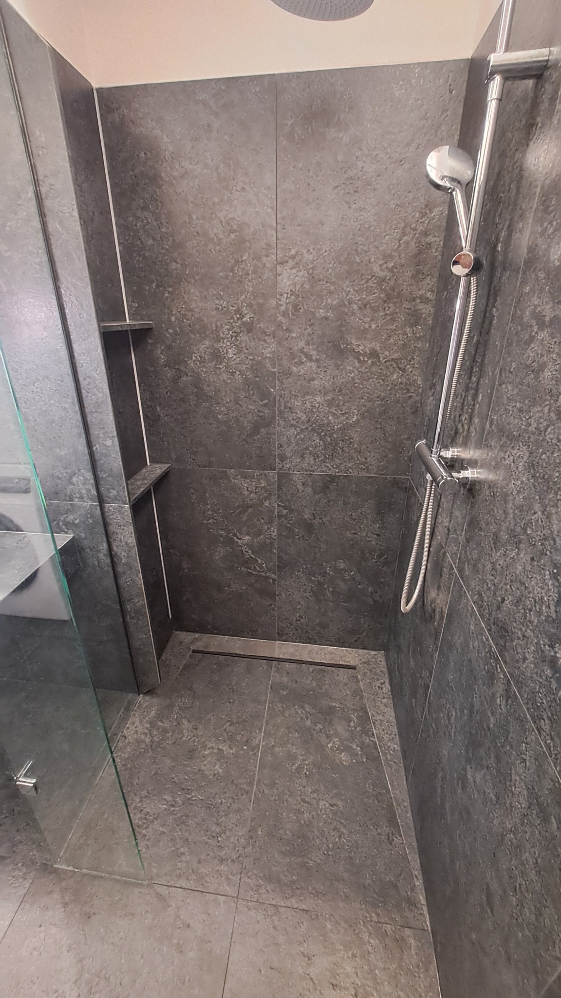 Nowoczesna kabina prysznicowa z szarymi płytkami imitującymi kamień, odpływ liniowy i półki wnękowe. Realizacja w Warszawie.