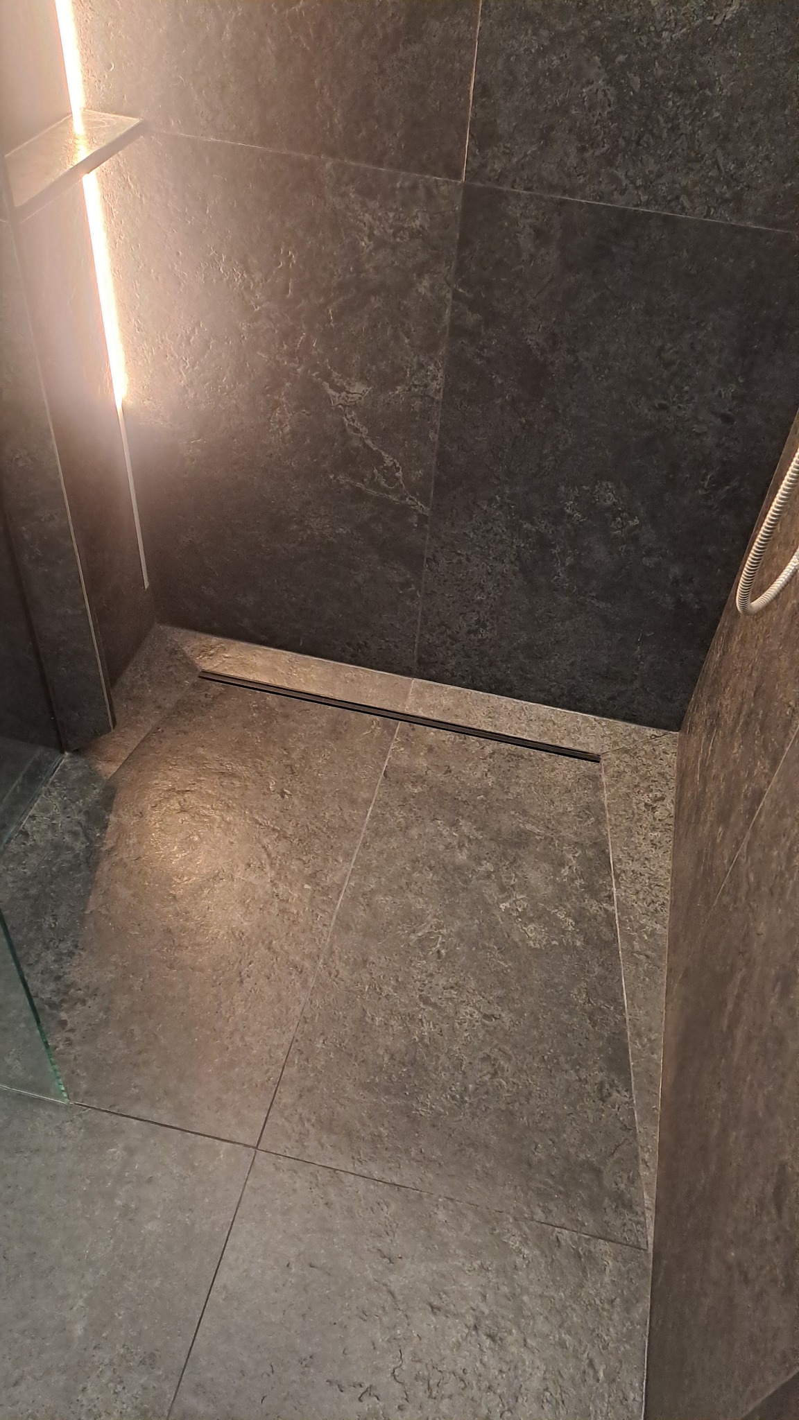 Nowoczesna łazienka z szarymi płytkami imitującymi kamień, czarnym odpływem liniowym i podświetlaną półką. Minimalistyczny design, eleganckie wykończenie.