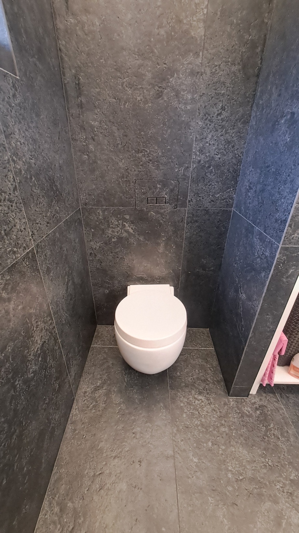 Nowoczesna toaleta podwieszana w minimalistycznej łazience wykończonej szarymi płytkami imitującymi kamień, z ukrytym systemem spłukiwania.