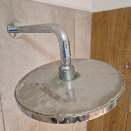 Serwis Sprzątający W Porządku - Zakamieniona, okrągła głowica prysznicowa z chromowaną armaturą, widoczne osady wapienne i zielonkawy nalot na łączeniu, na tle płytek ceramicznych i drewnopodobnej okładziny ściany.