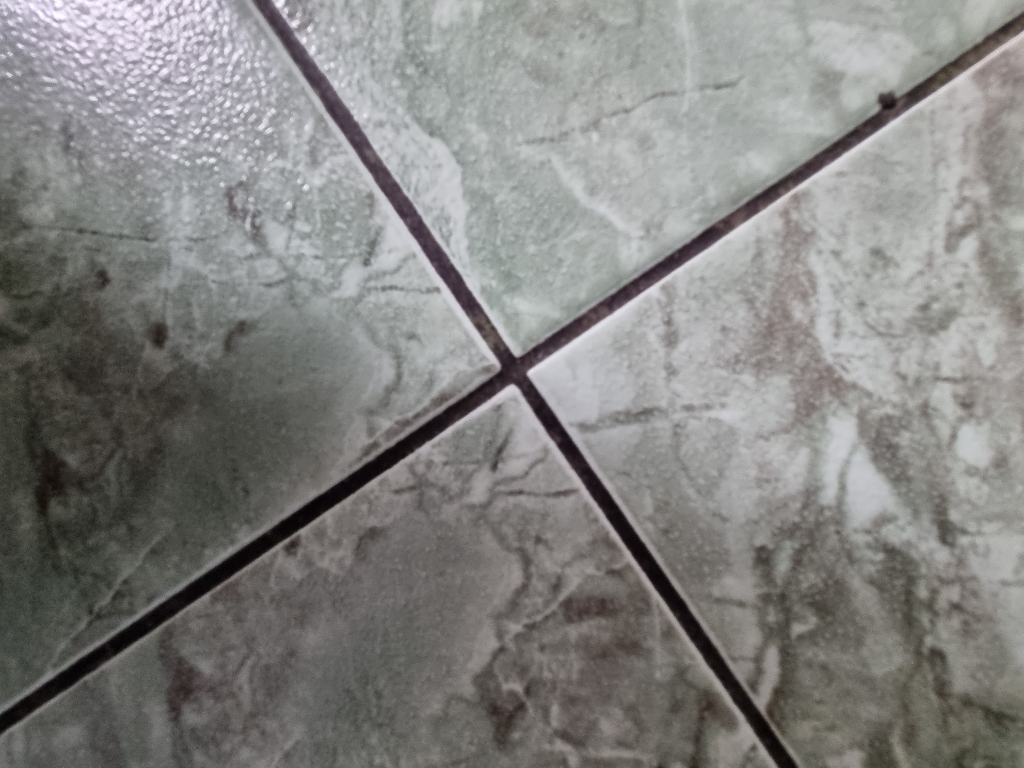 Szczegółowy widok na ułożone, błyszczące, zielonkawo-szare płytki podłogowe z ciemnymi fugami, tworzące krzyżujące się linie.