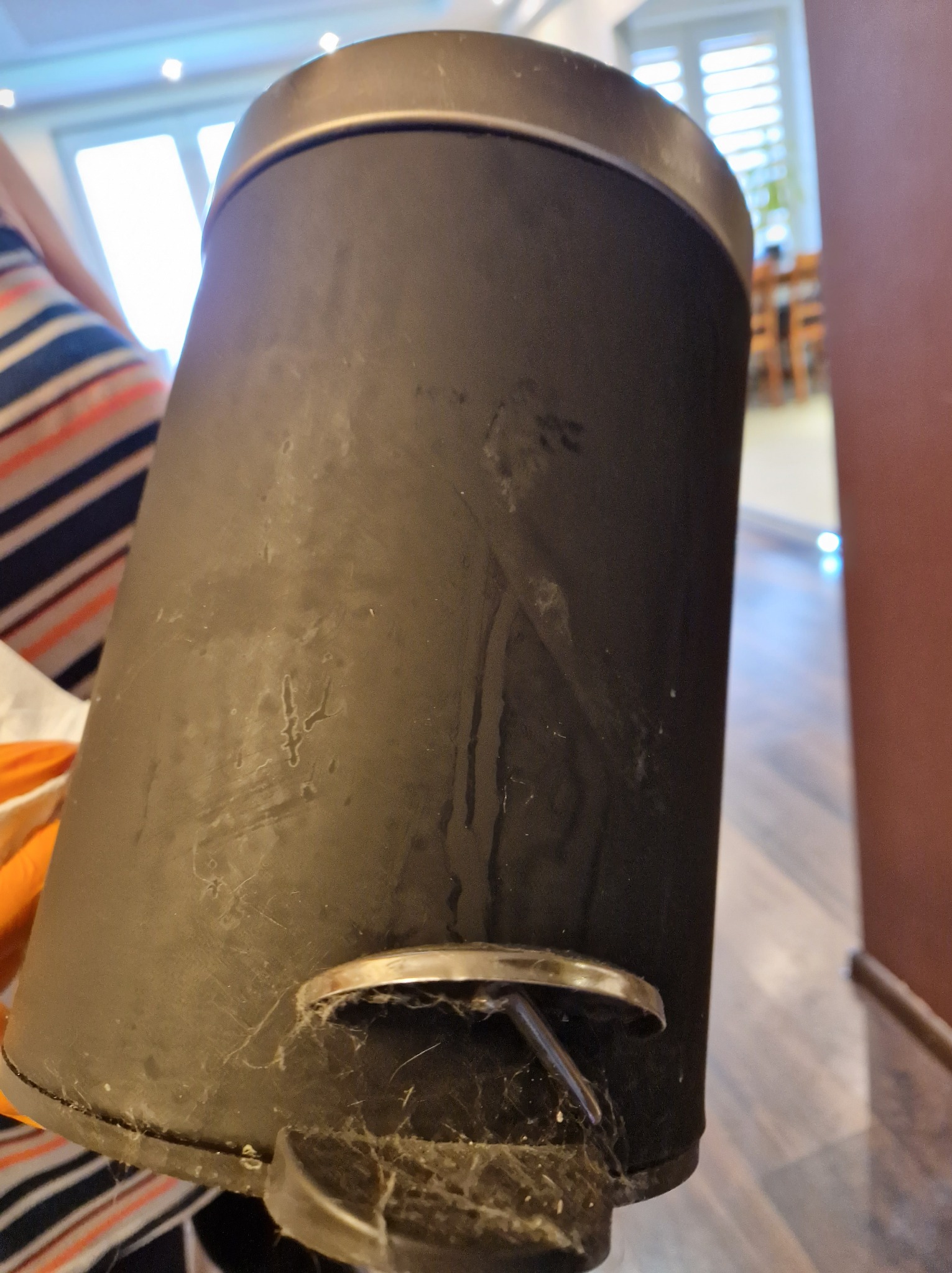 Brudny, czarny kosz na śmieci z pedałem, pokryty kurzem i pajęczynami, widoczne ślady użytkowania.