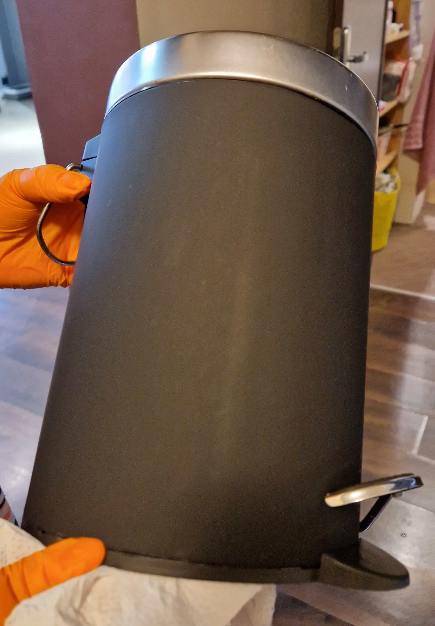 Czyszczenie matowego, czarnego kosza na śmieci z chromowanymi elementami. Ręka w pomarańczowej rękawicy czyści dolną krawędź kosza wilgotną szmatką.