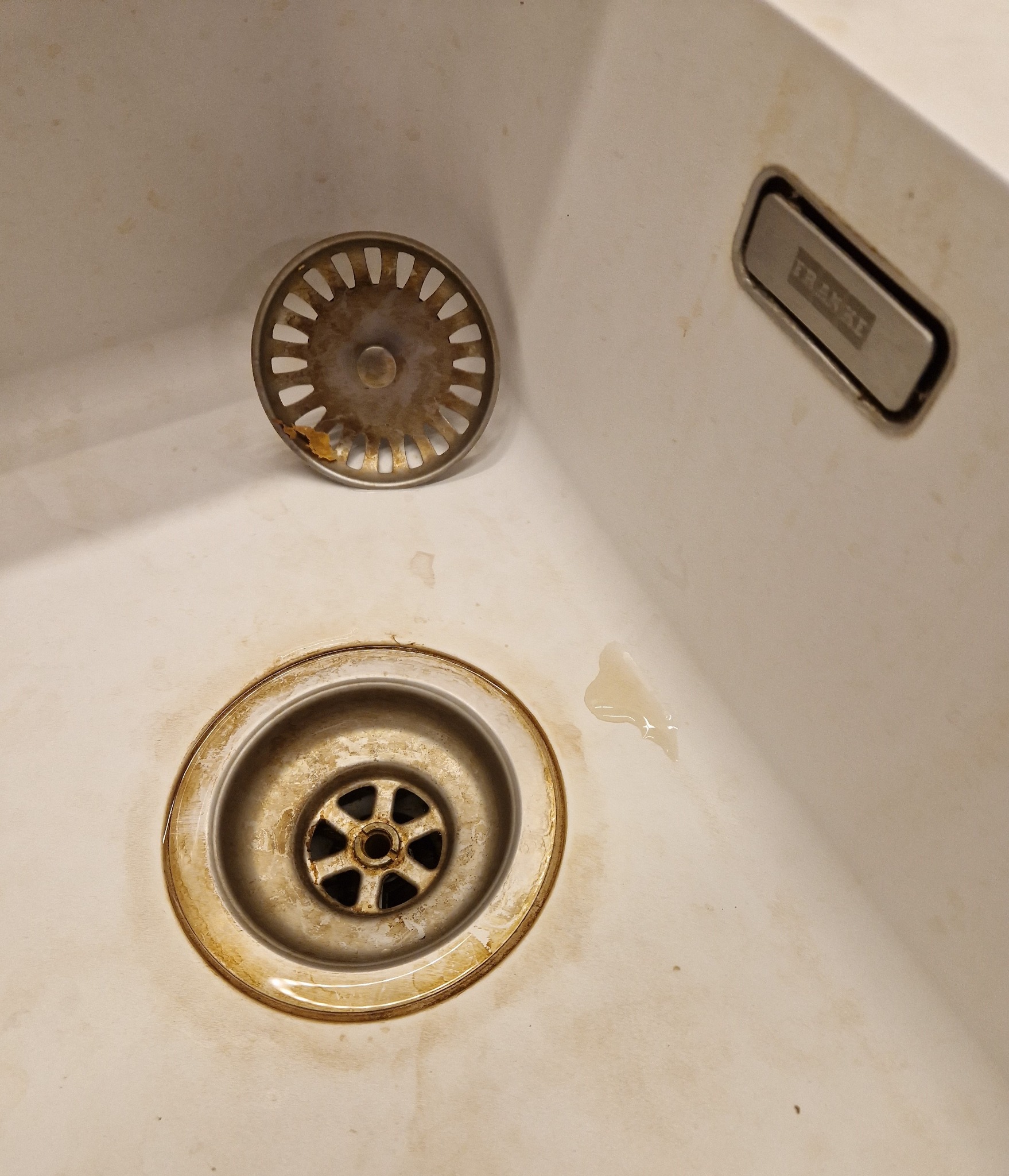 Zabrudzony zlew kuchenny z metalowym odpływem i wyjętym sitkiem, widoczne zacieki i osady kamienia, logo Franke w prawym górnym rogu.