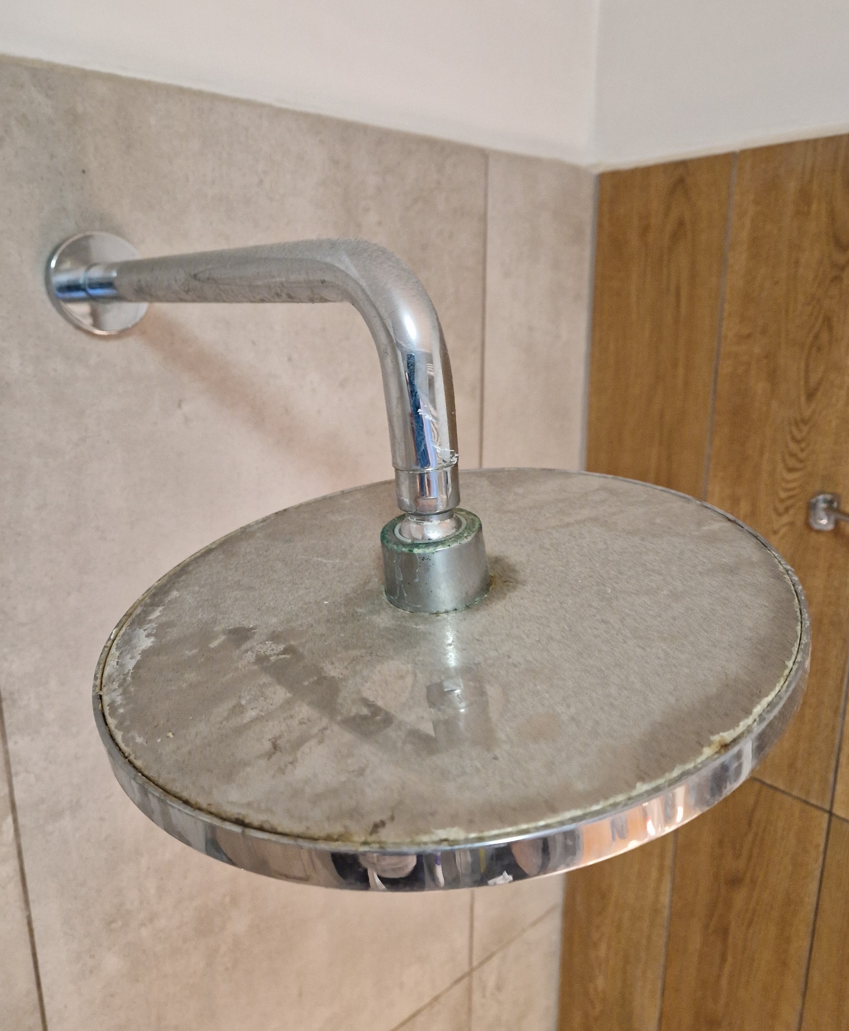 Zakamieniona, okrągła głowica prysznicowa z chromowaną armaturą, widoczne osady wapienne i zielonkawy nalot na łączeniu, na tle płytek ceramicznych i drewnopodobnej okładziny ściany.