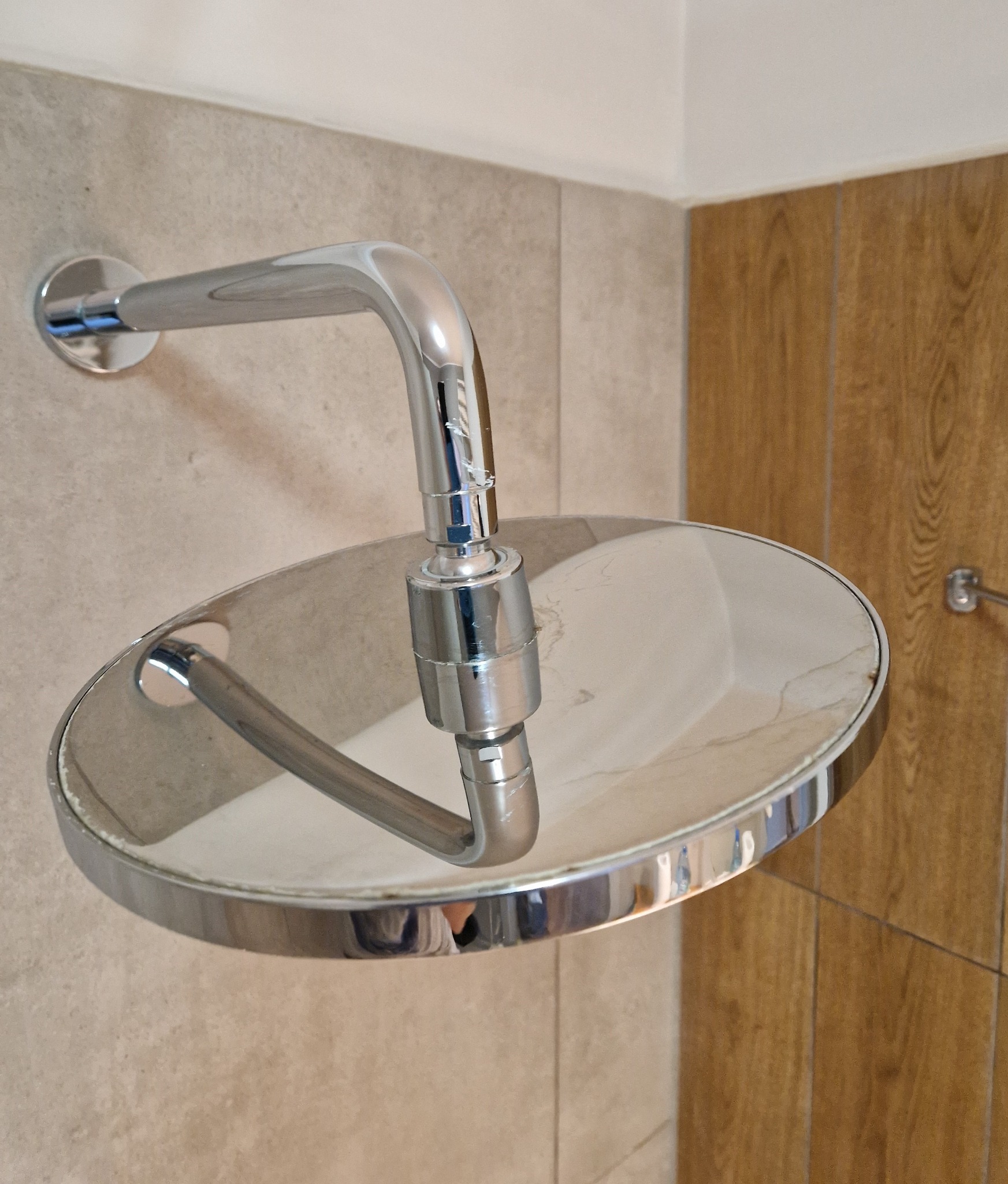 Błyszcząca, okrągła deszczownica chromowana zamontowana na ścianie wyłożonej beżowymi płytkami, z widocznymi zaciekami i śladami po wodzie na powierzchni.