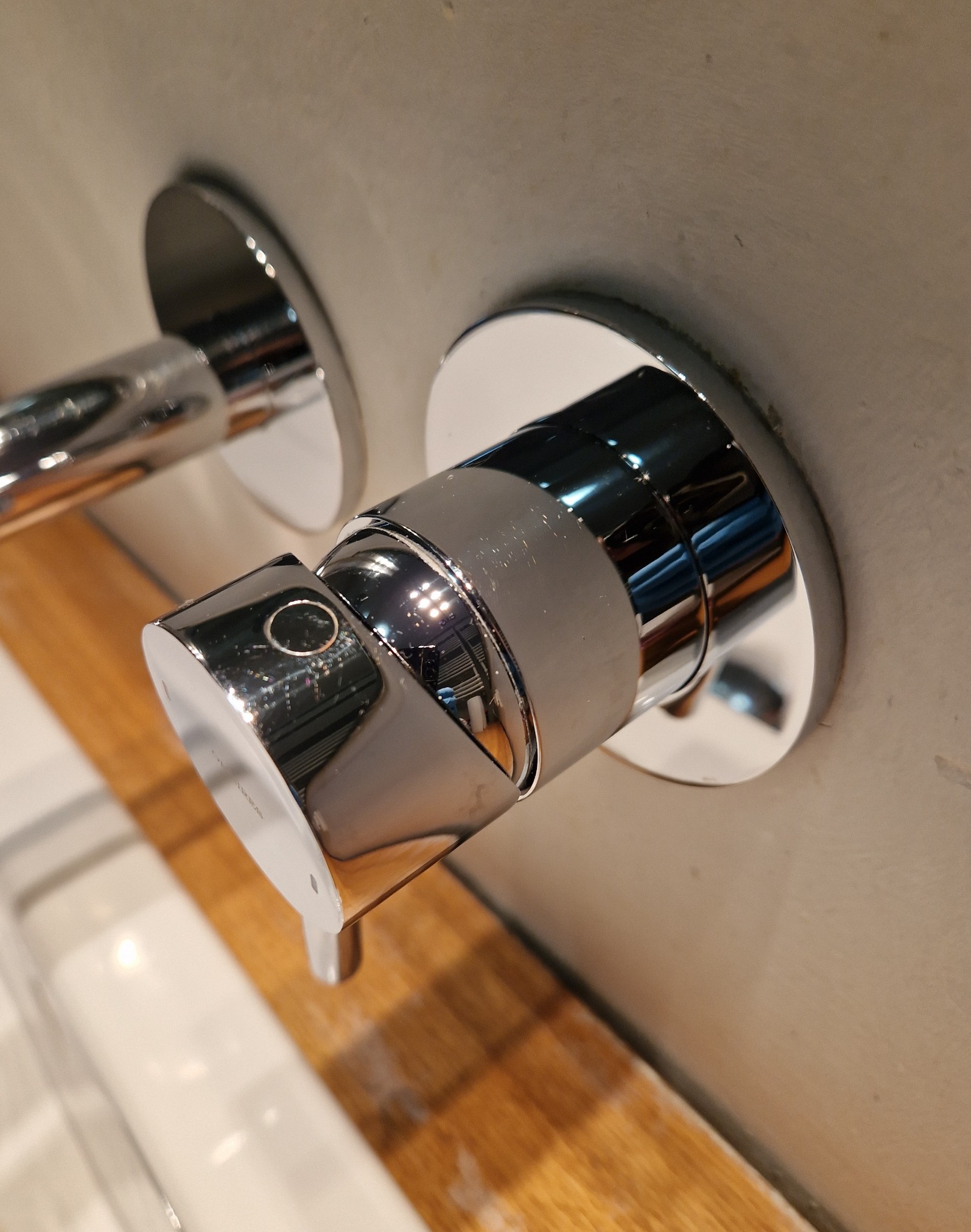 Błyszcząca chromowana bateria umywalkowa zamontowana na szarej ścianie, z widocznym fragmentem umywalki i blatu z jasnego drewna.