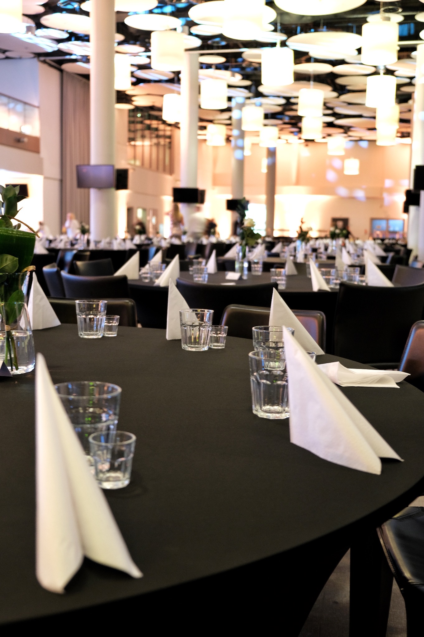 Elegancko nakryte stoły z czarnymi obrusami, szklankami i białymi serwetkami w sali bankietowej z dekoracyjnym oświetleniem sufitowym.
