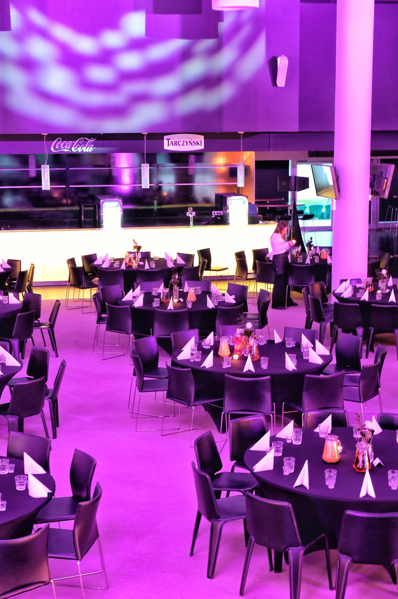 Sala bankietowa oświetlona fioletowym światłem, z okrągłymi stołami nakrytymi czarnymi obrusami, białymi serwetkami i szklankami, przygotowana do imprezy, w tle bar z logo Coca-Cola i Tarczynski...
