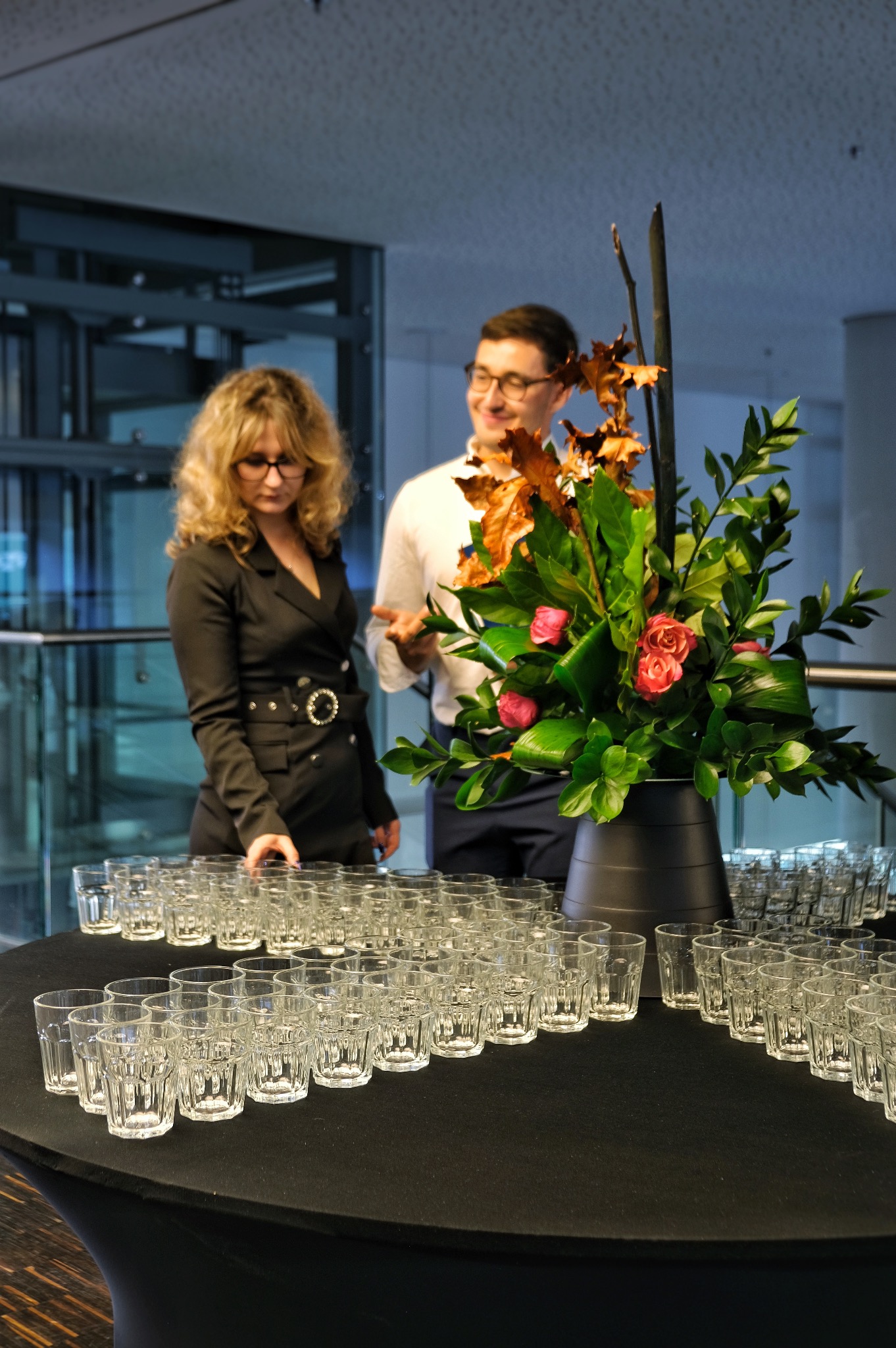Stół z czarnym obrusem, na którym ustawione są rzędy szklanek, w tle dekoracja kwiatowa i dwie osoby w eleganckich strojach, prawdopodobnie przygotowania do wydarzenia firmowego.