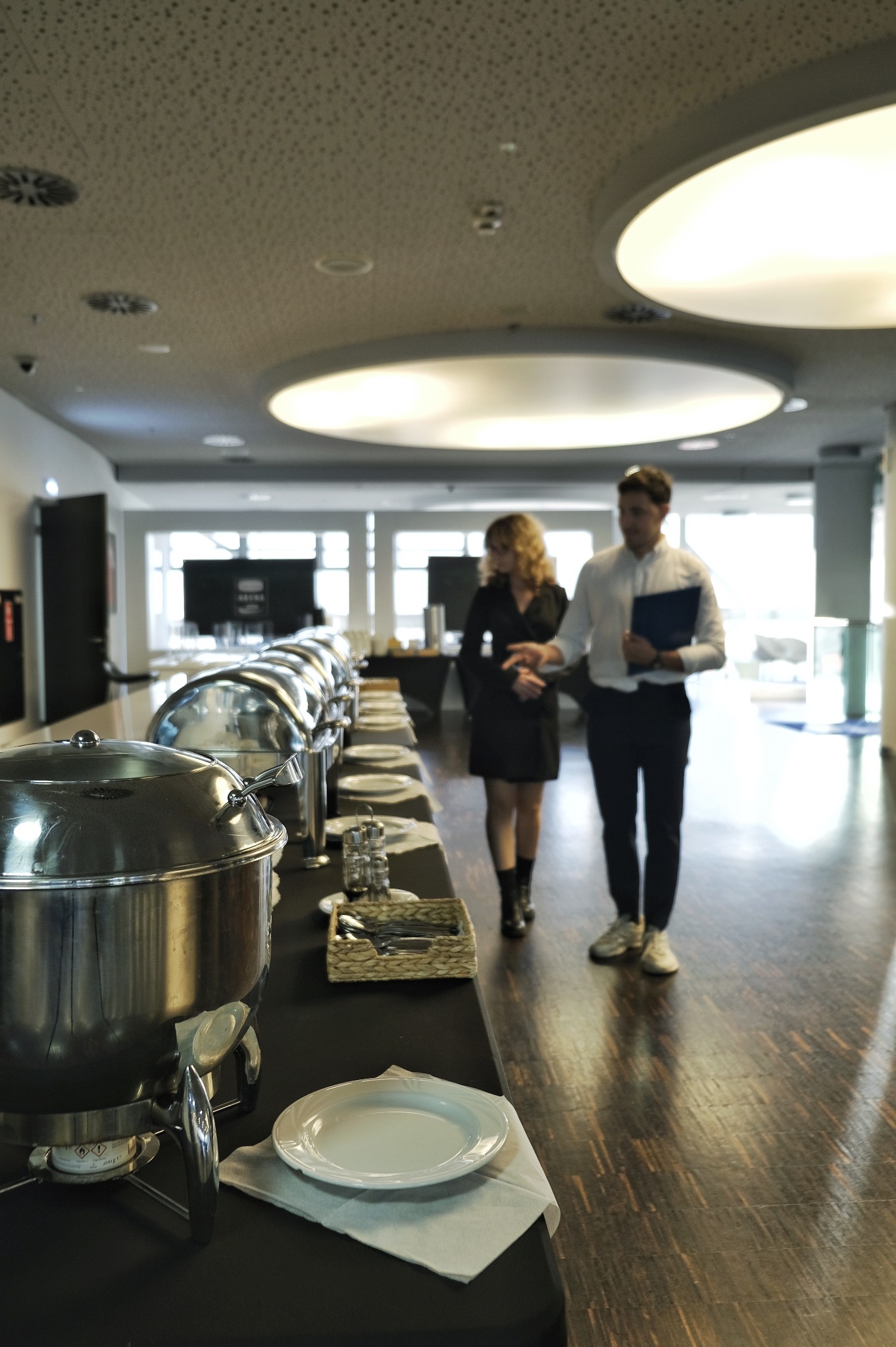 Długi stół z podgrzewaczami do potraw i zastawą, w tle para rozmawiająca w eleganckim wnętrzu z okrągłymi lampami sufitowymi.